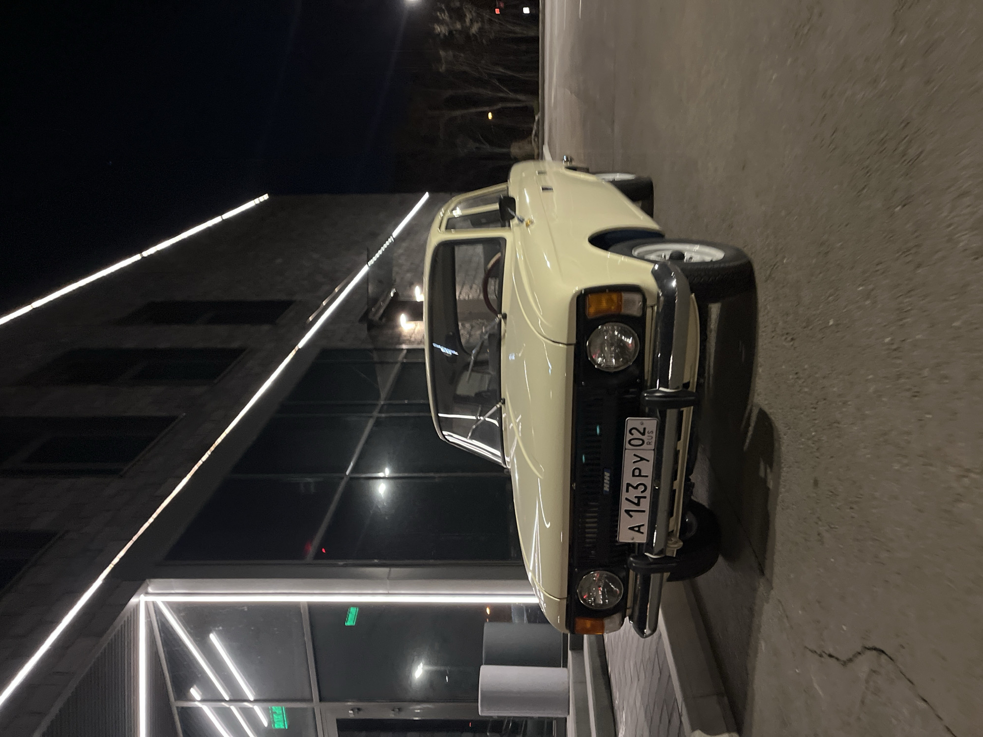 The image shows a light-colored vintage car parked on a paved surface next to a modern building with illuminated linear lighting. The license plate on the car reads "A 143 PY 02 RUS," indicating it is a Russian vehicle. The image was taken at night. The location is likely in Krasnoyarsk, Russia, given the license plate. There are no people visible in the image.