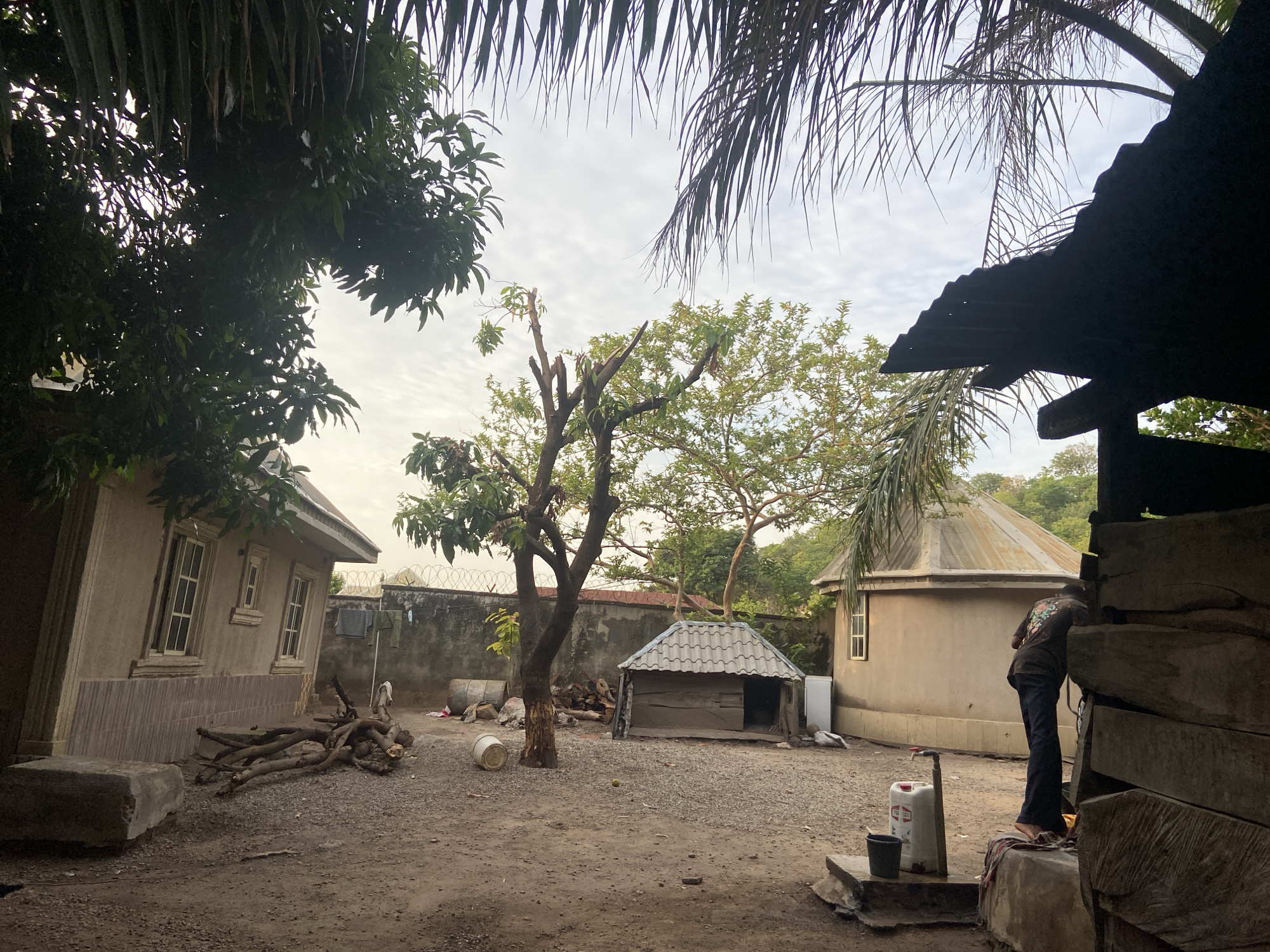 The image depicts a courtyard in Mararaba, Nigeria. The ground is dirt and gravel. There are two small buildings, one rectangular with a pitched roof and one with a conical roof, both seemingly made of stucco. A small wooden structure with a simple roof is positioned between the two buildings. A tall, leafless tree stands in the center of the courtyard. A man is standing near a water spigot, possibly washing something. There is laundry hanging on a barbed wire fence in the background. Branches and logs are piled near the rectangular building. A plastic jug and bucket are near the water spigot. Overhanging trees frame the upper portion of the image. The sky is overcast.