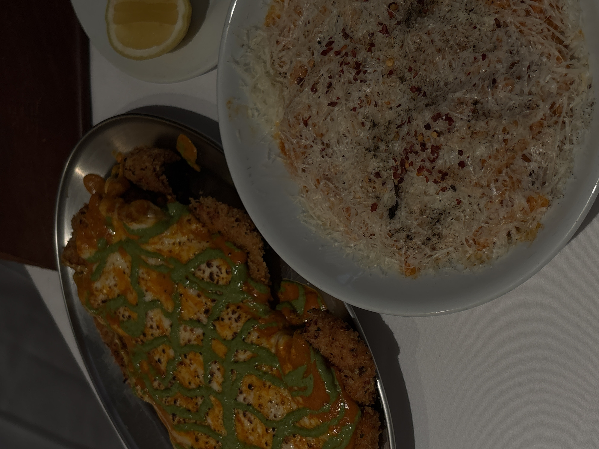 The image shows two dishes of food on a white tablecloth. The dish on the left appears to be a breaded cutlet topped with a red sauce and a green sauce drizzled in a crisscross pattern. The dish on the right is a bowl of pasta topped with grated cheese and red pepper flakes. A lemon wedge is on a small white plate to the right of the cutlet dish. The scene is illuminated by overhead lighting, casting shadows. The background suggests an indoor setting, possibly a restaurant. There are no people visible in the image, and no context is provided to suggest a location in Mexico City.