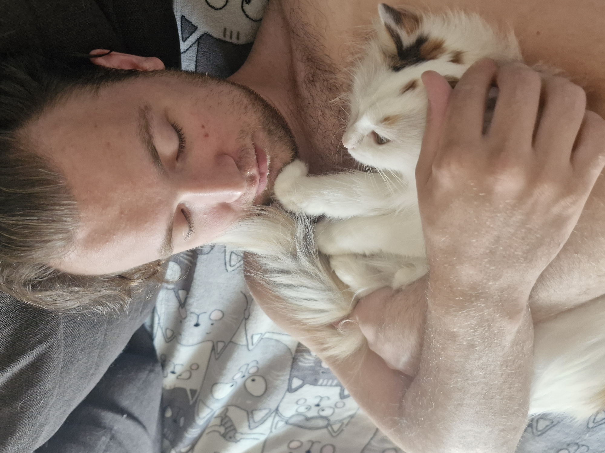 A man with closed eyes and light skin is lying down, holding a white cat with patches of brown and black fur. The cat is positioned against his chest. The man has short, wavy brown hair and facial hair. He is shirtless. The bed is covered with a grey and white patterned sheet. It is not possible to determine the location of this image as Yekaterinburg, Russia or any other city based on the provided information.
