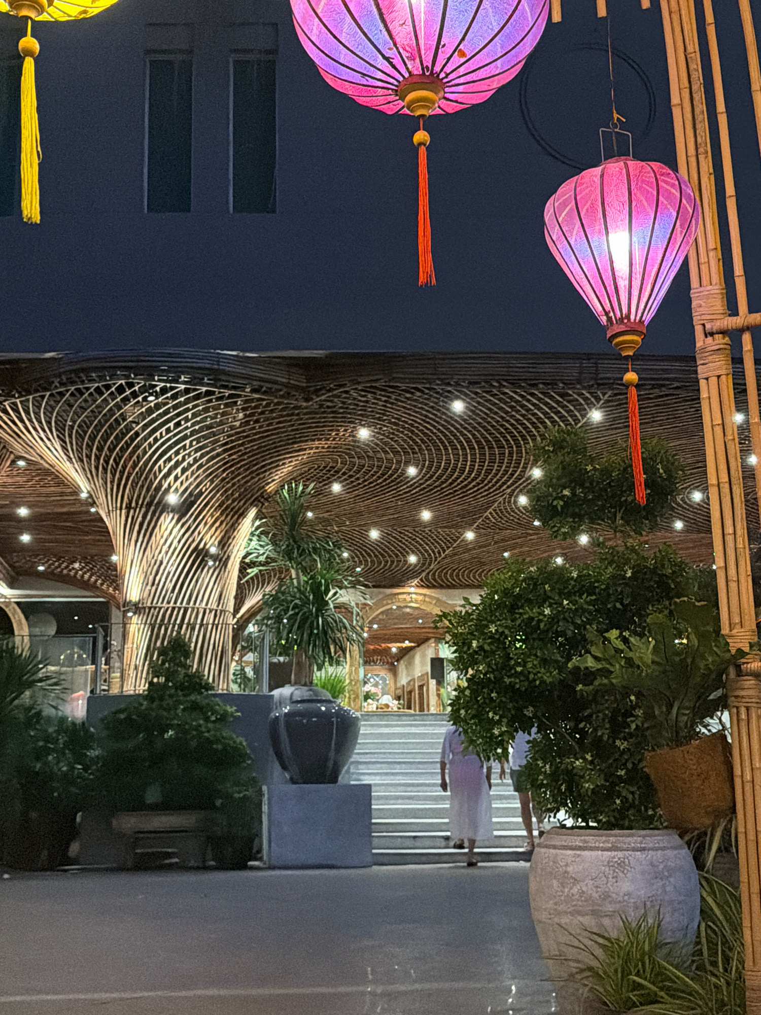 The image depicts an exterior scene at night, likely in Nha Trang, Vietnam, showcasing architectural elements and decorations. In the foreground, a large, ornate gray planter holds a lush green plant, with another smaller plant to its left. To the right, a bamboo structure stands, adorned with lighting. Above, three large, colorful lanterns are suspended; one is yellow, and two are shades of pink and purple. The lanterns have intricate patterns and tassels.
The middle ground features a grand staircase leading up to a building with a distinctive ceiling constructed of curved wooden beams and illuminated by numerous small lights, creating a swirling pattern. A dark, large vase sits on a pedestal to the left of the staircase. Two people are visible on the stairs, walking upwards; one is wearing a long white garment, and the other is wearing shorts and a light-colored shirt.
The background reveals the upper facade of the building, which is dark and features two rectangular windows. The overall atmosphere is festive and illuminated, suggesting a public space or a venue decorated for an event. The presence of bamboo and the style of the lanterns are characteristic of Southeast Asian aesthetics.