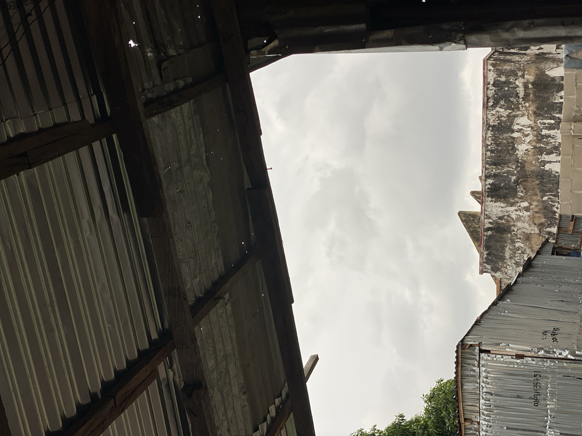 This is a photograph taken in Jalingo, Nigeria, from beneath a corrugated metal roof supported by wooden beams. The roof shows signs of wear, with some holes visible. Outside, the sky is overcast with gray clouds. In the mid-ground, there is a corrugated metal structure with writing on it, and beyond that, a damaged building with peeling plaster. There is also a glimpse of green foliage from a tree on the left side of the image. The overall impression is one of a somewhat weathered and utilitarian urban environment.