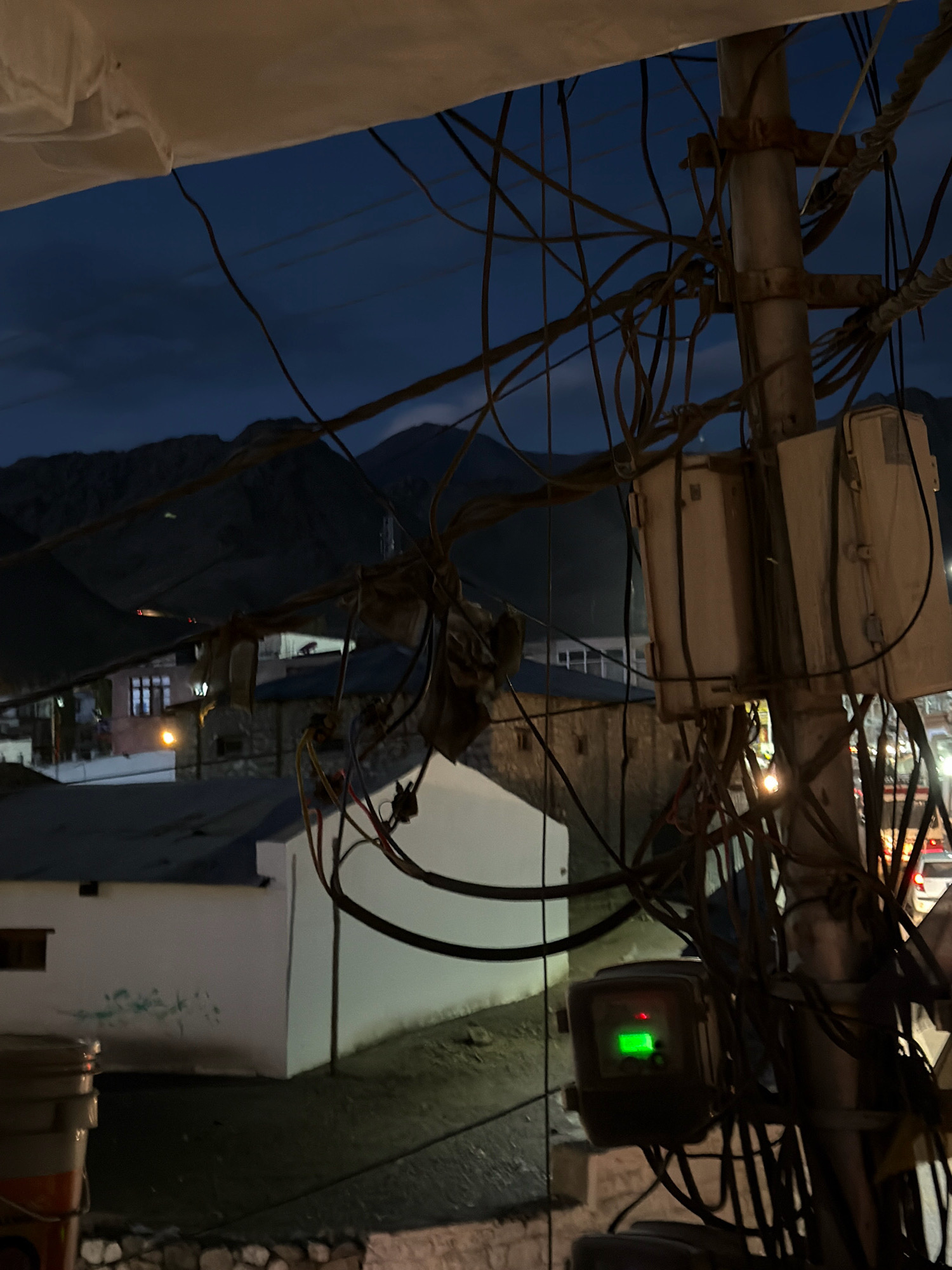 The image captures a scene at dusk in Leh, India, with a focus on a utility pole heavily laden with electrical wires and several gray, rectangular junction boxes. The pole itself is metallic and appears weathered. Wires of various thicknesses crisscross the frame, some bundled together and others hanging loosely. One junction box has a small, glowing green light on its surface. In the background, the dim light of evening illuminates a cluster of buildings with flat roofs and some windows lit from within. Beyond the buildings, dark, mountainous silhouettes are visible against the twilight sky, which is a deep blue. The overall impression is one of a utilitarian, somewhat cluttered urban environment at the close of day.