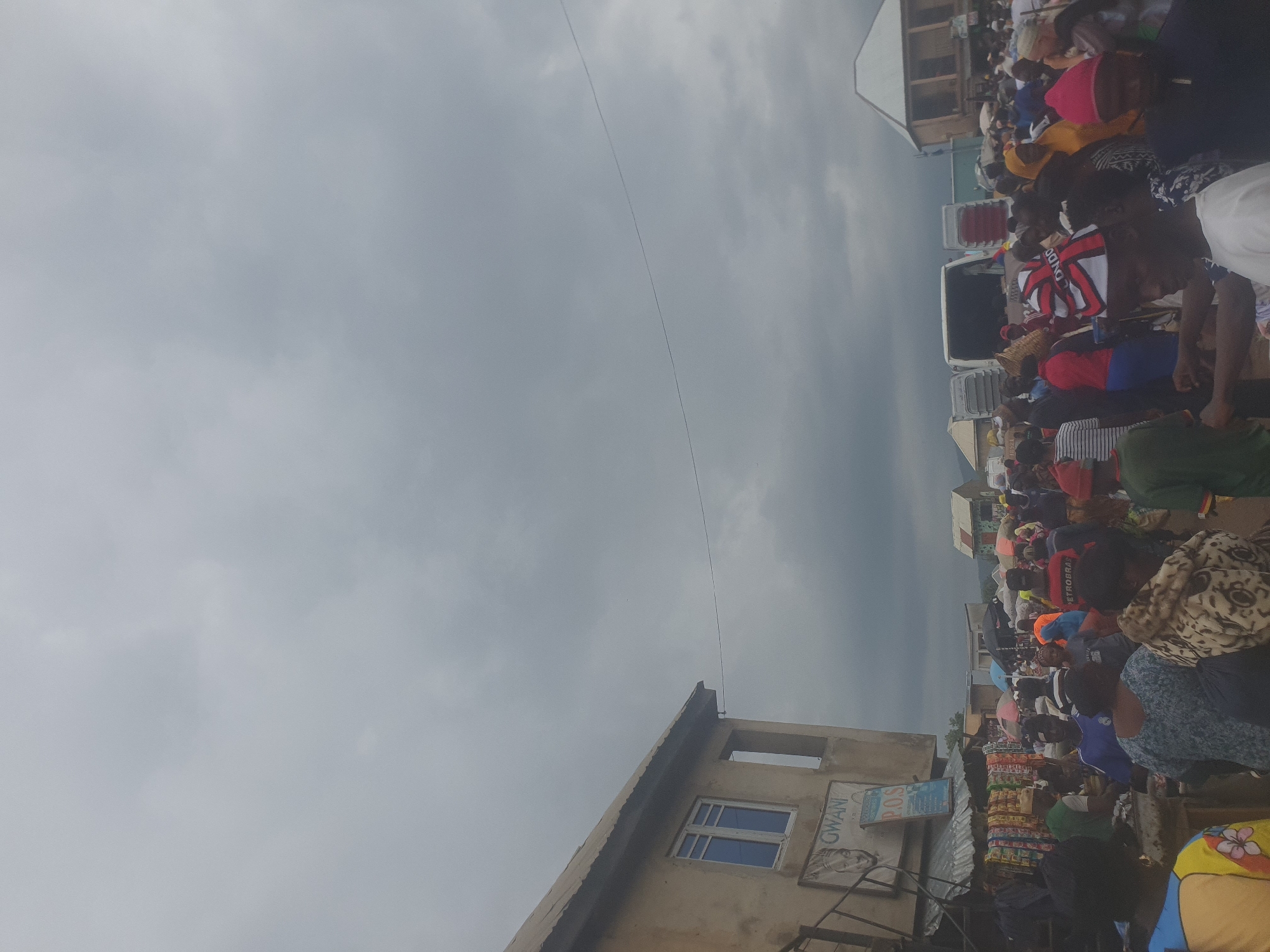 This media file depicts a bustling outdoor scene in Rafin Par, Nigeria, likely a market or gathering point. A large crowd of people, men and women of various ages, are densely packed together. Many are dressed in colorful clothing. Some are engaged in what appears to be shopping or waiting. Objects visible include numerous market stalls laden with goods, predominantly food items and packaged products. There are also several vehicles, possibly buses or vans, suggesting transportation or logistics. A building with windows and signage is partially visible, indicating an urban or semi-urban environment.
The activity appears to be a lively commercial exchange or a community event. The mood seems active and engaged, with people interacting and moving within the crowded space. The weather appears overcast, with a grey, cloudy sky, suggesting it might be late afternoon or a generally cloudy day.
Visible text includes "GWANI" and "P.O.S." on signage. The overall impression is one of vibrant local life and commerce in Nigeria.