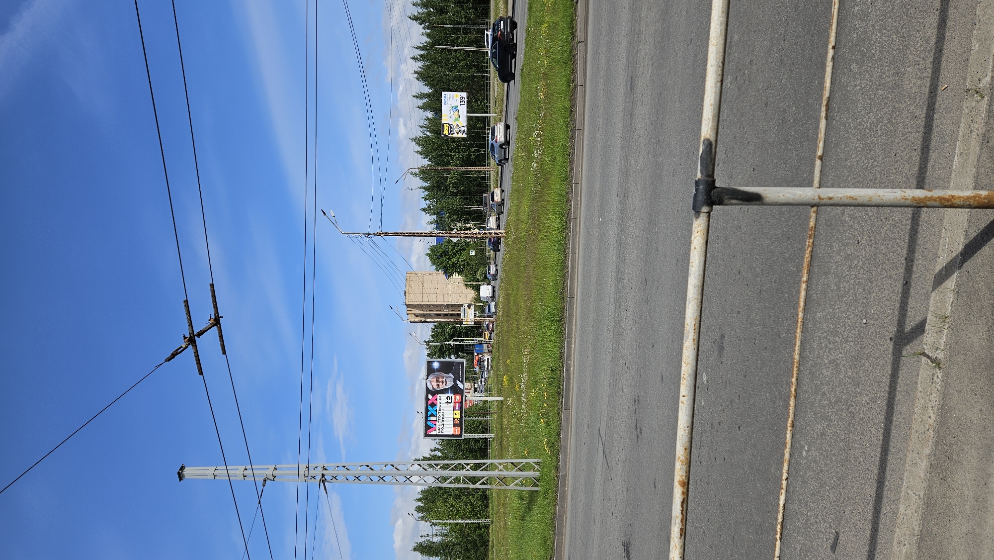 The image captures a street scene in an urban environment, likely in the city of Unknown, Unknown. The foreground features a section of a rusted metal barrier running parallel to the curb of an asphalt roadway. Adjacent to the roadway is a strip of green grass, interspersed with patches of yellow wildflowers. Beyond the grass strip, a line of trees forms a green backdrop. In the distance, vehicular traffic is visible on the road, including cars and trucks. Several billboards can be seen along the roadside, displaying advertisements. Overhead, electrical power lines stretch across the clear blue sky.