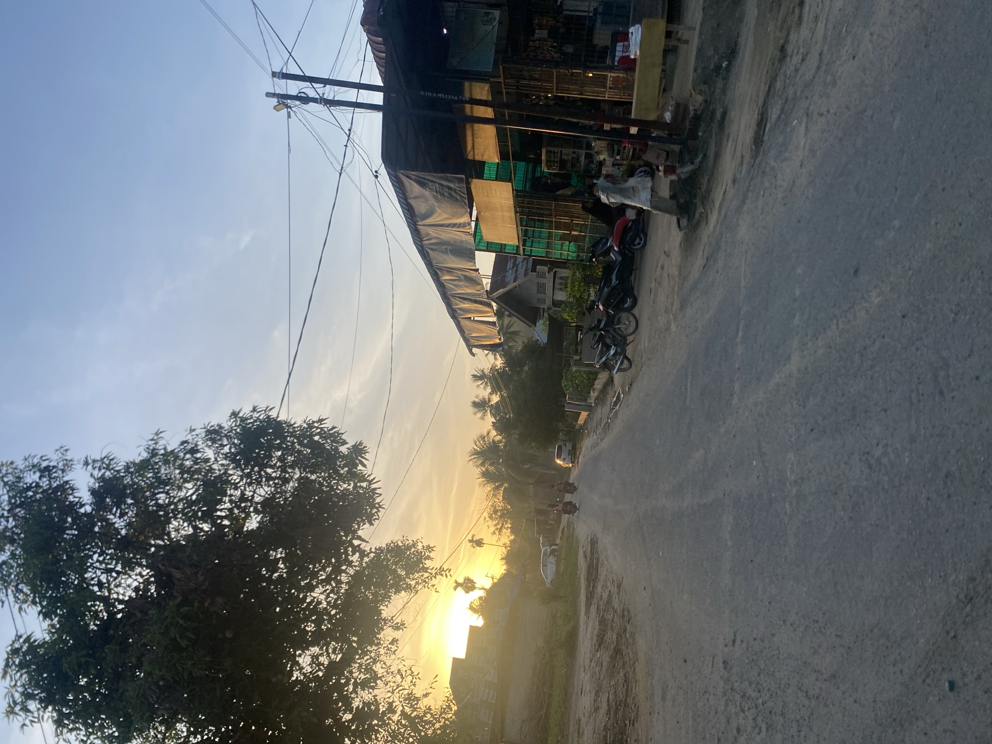 The image depicts a road in Kubu, Indonesia, during sunset. The road runs into the distance towards the setting sun, with buildings and vegetation on either side. A large tree is visible on the left, partially obscuring the view. Power lines are strung overhead. Parked motorcycles are located near a building on the right side of the road. The road surface is asphalt. Further down the road, there are vehicles and motorbikes driving in the distance.