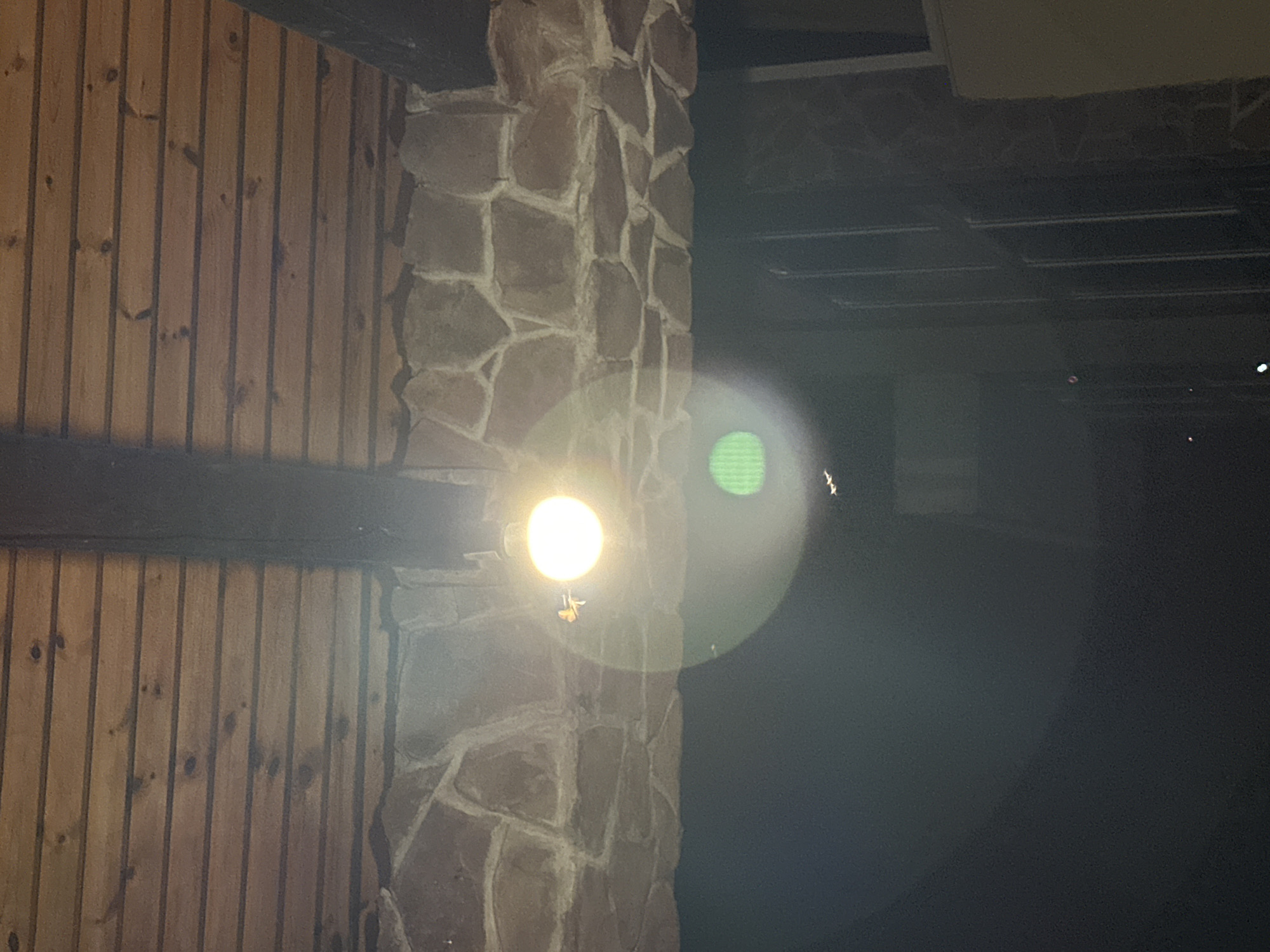 The image displays a close-up view of a porch ceiling constructed from horizontal wooden planks and a stone-clad overhang. A single, brightly lit bulb illuminates the scene, causing a significant lens flare that creates a circular green haze and a fainter rainbow effect. A small insect, possibly a moth, is visible in the vicinity of the light bulb. The dark area below the overhang suggests it is nighttime or an indoor setting with minimal ambient light. No specific landmarks or details placing the image in Sochi, Russia, are present within the frame.