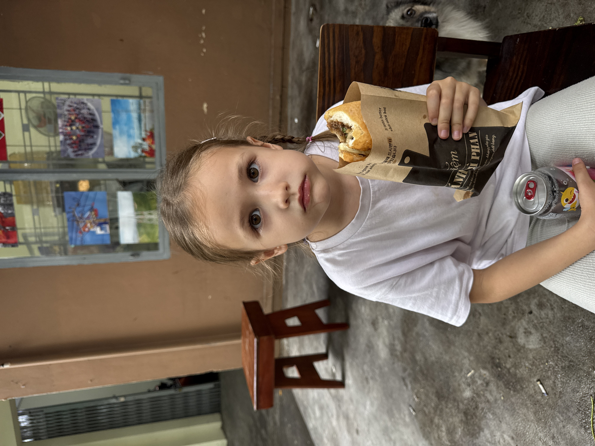 A young girl sits indoors, holding a partially eaten sandwich in a brown paper bag labeled "Nệm NHẬT PHAN," and a can decorated with a cartoon shark. She is wearing a white t-shirt and has her hair in braids. A small wooden stool sits on the concrete floor behind her. To her left, a window displays various items including what appear to be pictures. The background features a brown wall, and a dog's head is visible behind the chair she is sitting on. Text in Vietnamese and Korean on the bag indicates the location in Nha Trang Ward, Vietnam.