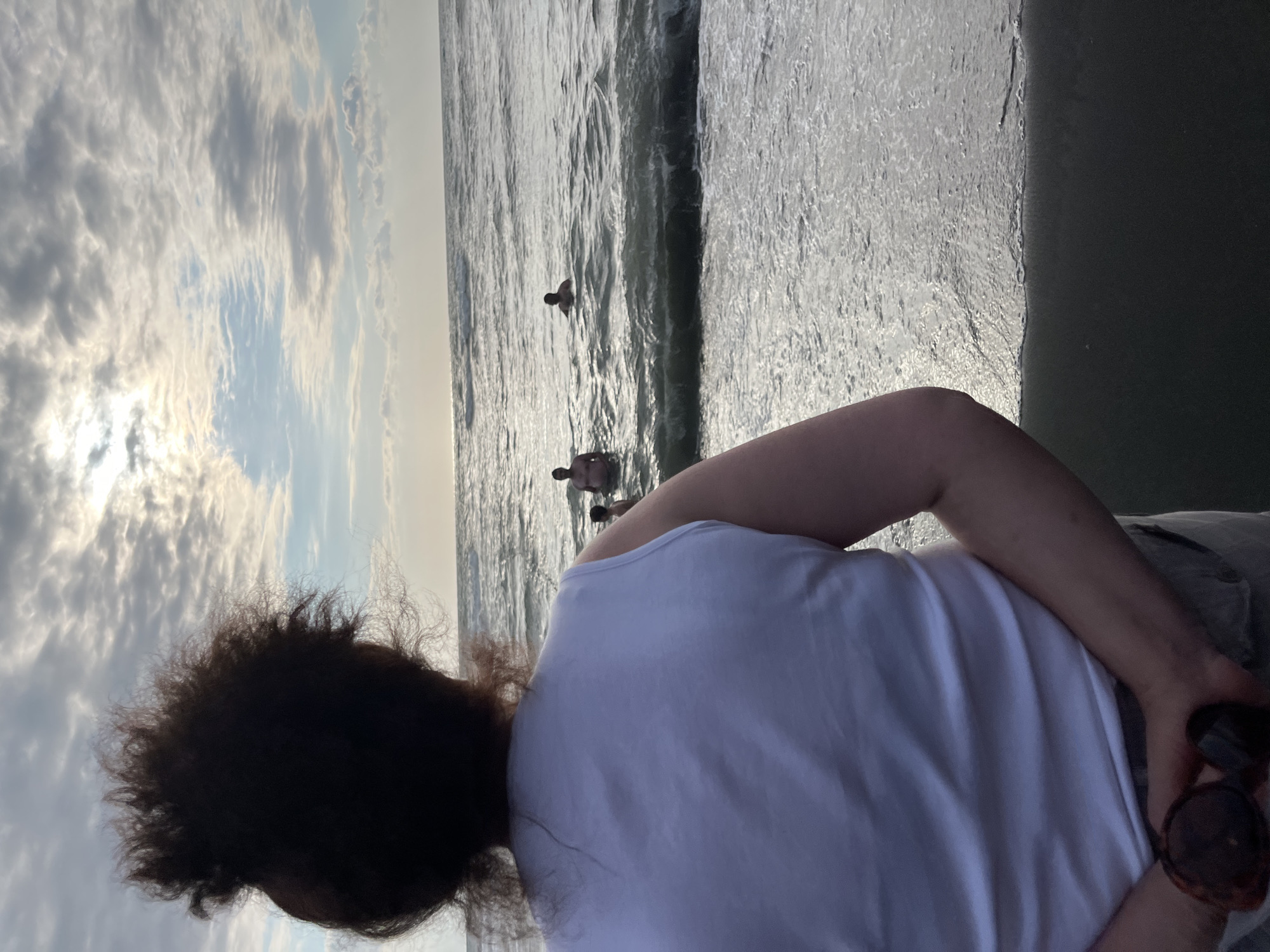 An overhead shot shows a person facing the ocean, with their back to the camera. The person has dark, curly hair and is wearing a white sleeveless shirt and light-colored pants. They are holding a pair of sunglasses in their hands, clasped behind their back. In the distance, three individuals are visible in the ocean. The sky is overcast with clouds, and the sun is peeking through, casting a bright glare on the water. The scene is the beach in Ureki, Georgia, known for its magnetic black sand.