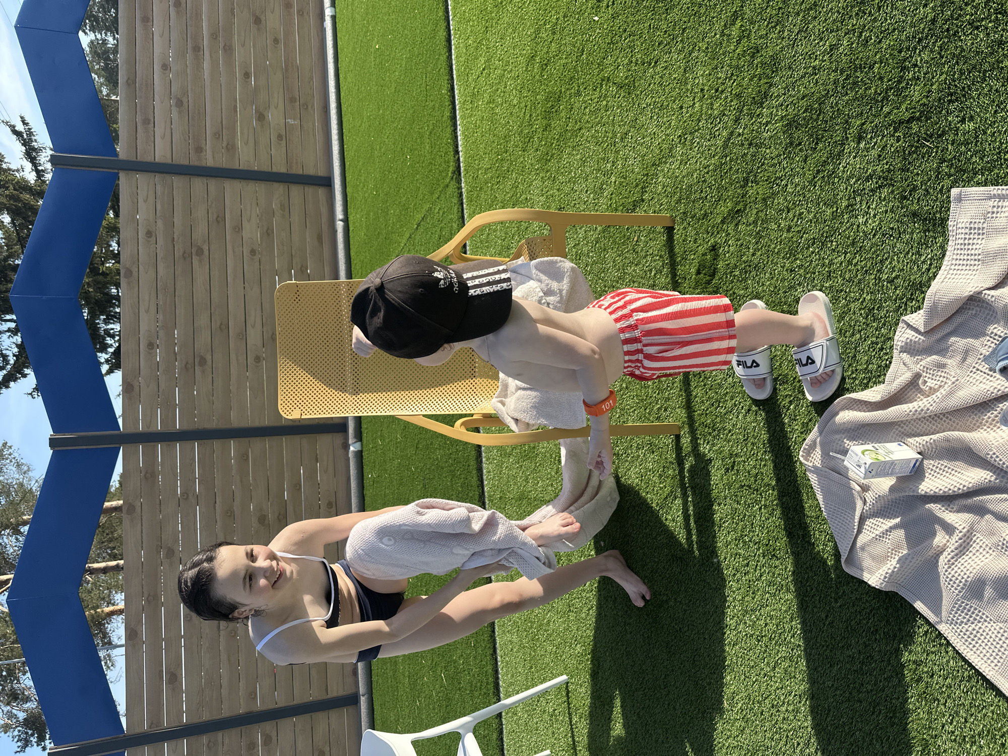 Scene: An outdoor scene in Votkinsk, Russia featuring two individuals near a swimming pool.
People: A young woman is seated on a white chair. She is wearing a black and white swimsuit and is drying someone's feet with a light-colored towel. A child wearing red and white striped shorts, white FILA-branded sandals, and a black baseball cap is standing on a yellow chair. They are also being dried off with a light-colored towel.
Objects: The scene includes a yellow metal chair, a white plastic chair, light-colored towels, a closed juice or milk container on a checkered towel.
Environment: The setting appears to be a backyard or pool area with artificial green grass. A fence made of vertical wooden planks with a blue canopy is in the background. The lighting suggests it is daytime.