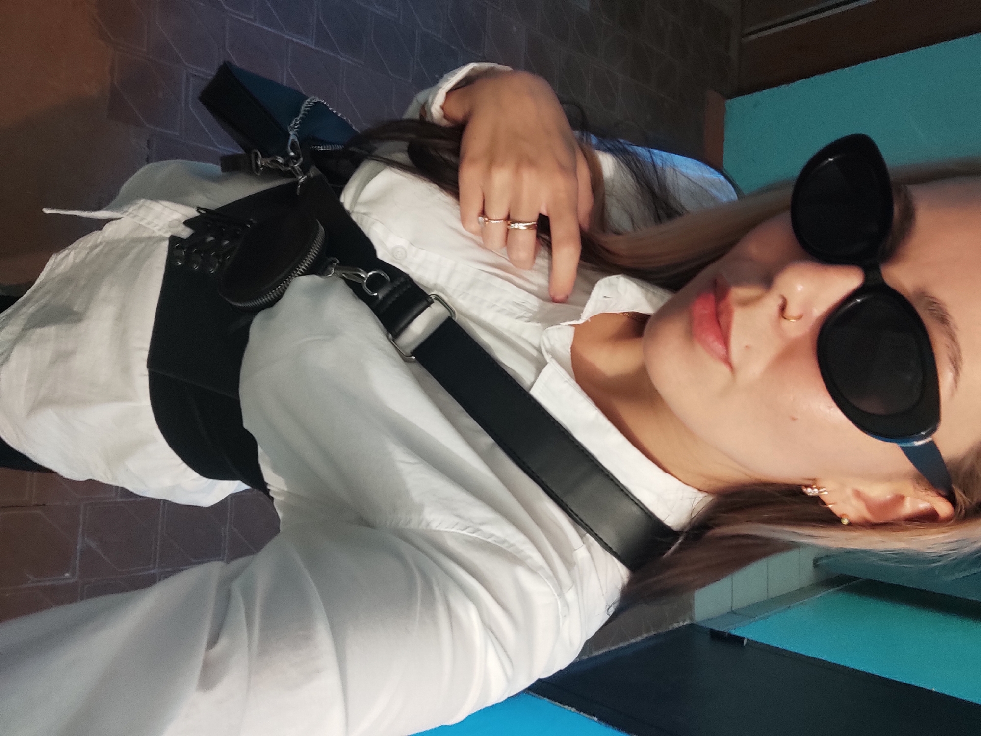 A selfie of a woman wearing a white button-down shirt and black sunglasses. She has a black leather harness with a small pouch over the shirt. She is wearing rings on her fingers and an earring in her left ear. The background is partially visible, showing a blue wall and a dark brown floor. It is impossible to verify that the photo was taken in the city of Chaikovsky, Russia.