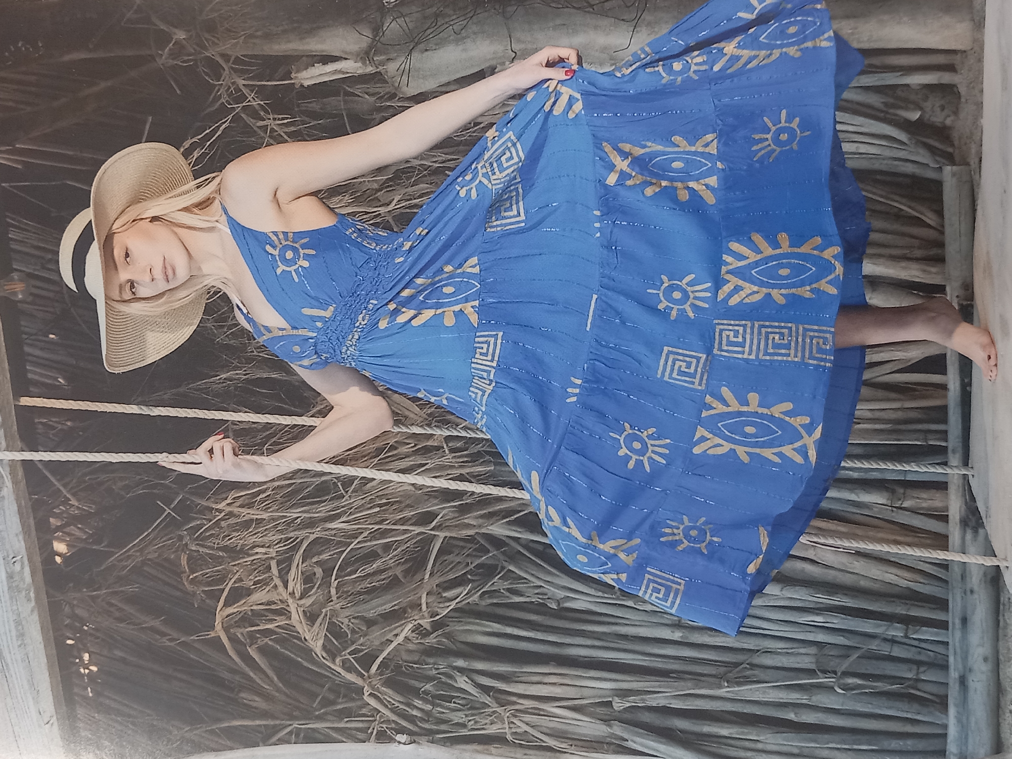 A blonde woman wearing a wide-brimmed straw hat and a blue tiered dress with gold patterns stands on a swing. The woman is on the right side of the frame, looking at the camera with a serious expression. Her right hand is on the rope of the swing, and her left hand is holding the hem of her dress. The swing is suspended from a wooden structure, and behind the woman is a wall of dried reeds or palm fronds. The ground beneath her is a light-colored wooden surface. The overall scene suggests an outdoor, possibly tropical, setting.