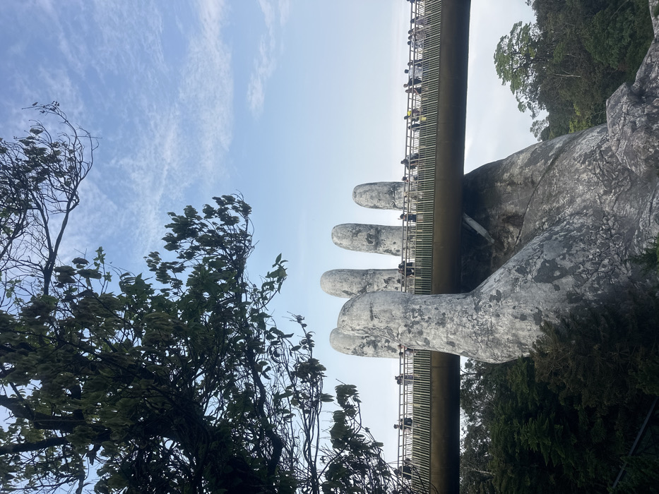 The image displays the Golden Bridge (Cầu Vàng) located in Ba Na Hills, Hòa Ninh, Hòa Vang, Đà Nẵng, Vietnam. The elevated pedestrian bridge, characterized by a golden-yellow pathway and a light-colored railing with numerous vertical supports, is prominently featured. It is visibly supported from below by two massive, weathered, faux-stone hands appearing grey and textured. One hand is fully visible in the foreground, extending upwards from a forested slope. Portions of fingers from a second hand are visible further along the bridge to the right.
A significant number of individuals are present on the bridge, distributed along its visible length, engaging in walking and observation. Their attire appears casual.
The environment includes a clear to partly cloudy blue sky with scattered cirrus clouds. Lush green trees and dense foliage are visible below and surrounding the bridge, indicating a mountainous or heavily vegetated setting. A large tree with dark green leaves and some bare branches occupies the upper left quadrant of the frame, partially obscuring the sky and upper portion of the bridge. The scene is illuminated by bright, natural daylight.