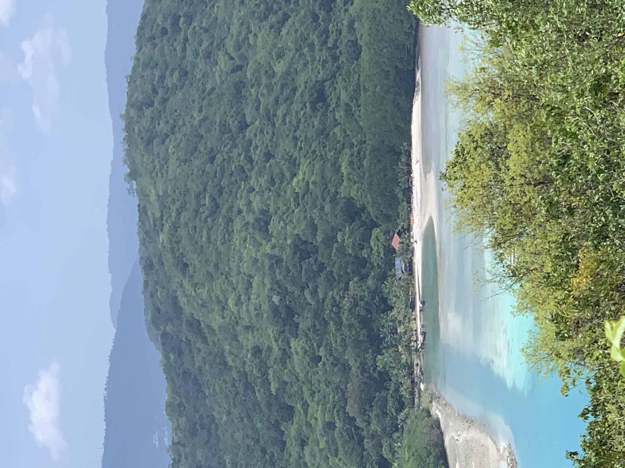The image depicts a tropical coastal scene. In the foreground, lush green foliage obscures the lower portion of the frame. Beyond the foliage, a body of clear, turquoise water stretches towards a sandy beach. Several small boats are anchored in the water near the shore. Along the beach, there are a few buildings, some with red roofs and solar panels, nestled amongst palm trees. The middle ground is dominated by a densely forested, green hillside that rises steeply. In the background, layers of hazy blue mountains recede into the distance under a pale blue sky with a single, fluffy white cloud. The scene is one of natural beauty and apparent tranquility. The location is in an unknown city in Thailand.