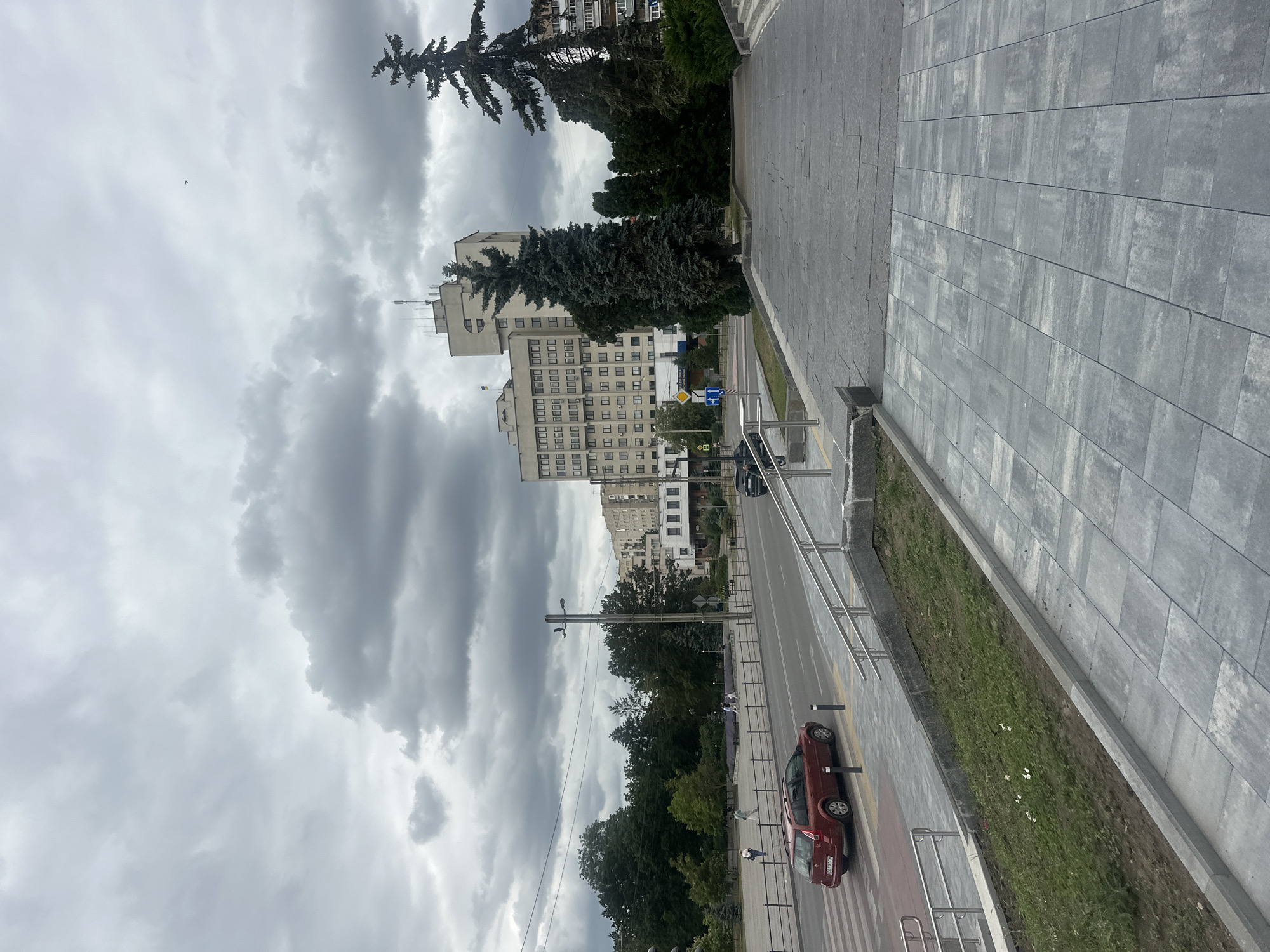 This image captures a street scene in Ternopil, Ukraine, under an overcast sky. The foreground is dominated by a wide, paved walkway with a textured grey stone tile pattern. A grassy area with some sparse white flowers is visible to the left of the walkway, bordered by a concrete curb and metal railings.
Beyond the walkway, a street runs horizontally across the frame. A red station wagon is stopped at a pedestrian crossing. Further down the street, several people are walking on a paved area next to a line of trees and bollards. A dark-colored vehicle is parked on the right side of the street.
In the background, a large, multi-story building with a light-colored facade and numerous windows stands prominently. Smaller buildings and more trees are visible on either side. Two tall evergreen trees are positioned in front of the buildings. The sky is filled with heavy, grey clouds, suggesting a cloudy or potentially rainy day. The overall impression is of an urban environment with pedestrian areas, a road, and commercial or administrative buildings.