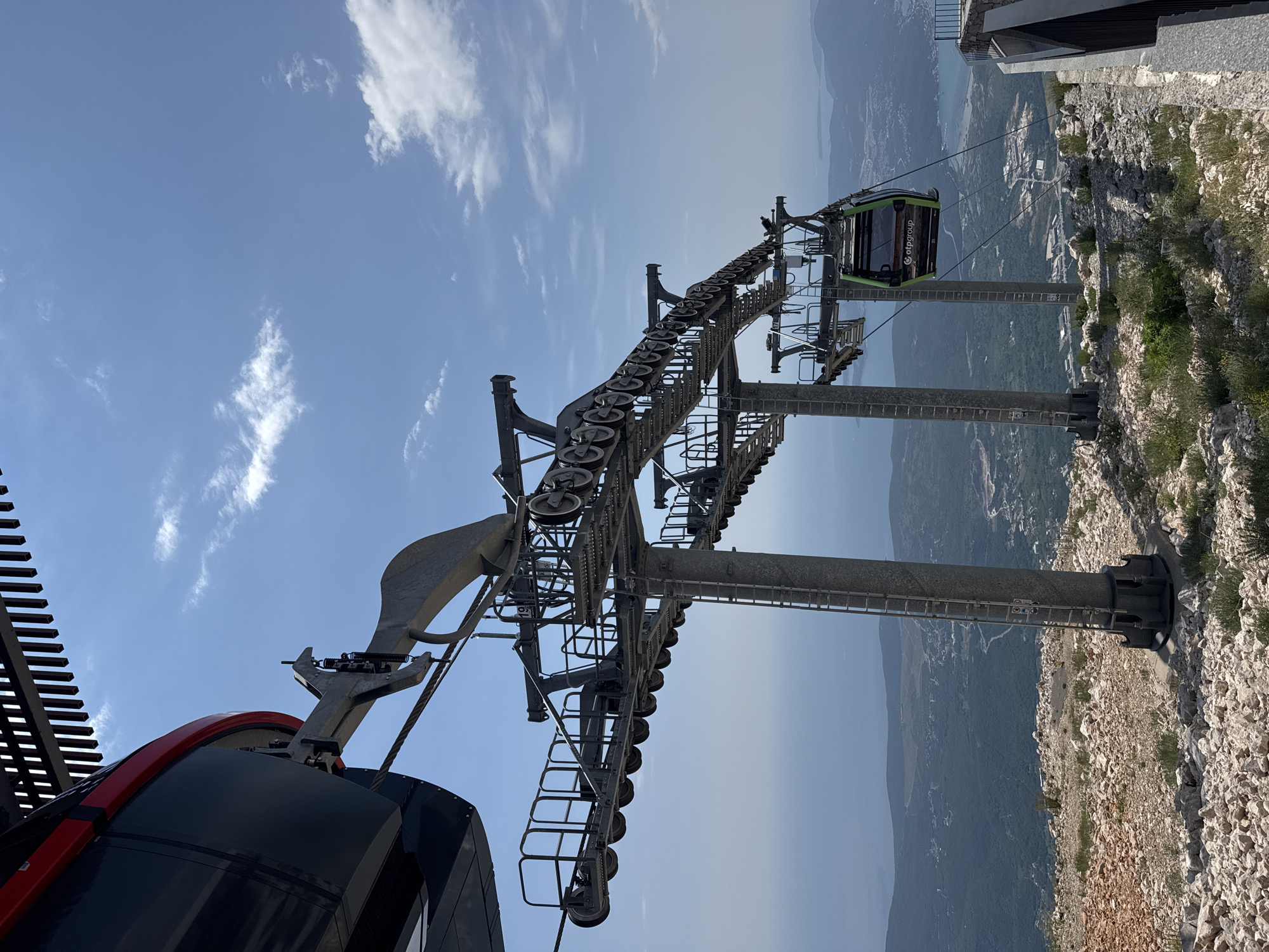 A ski lift is visible at the top of a mountain with a view of the sea and a town in the distance. The ski lift is made of metal and has several cars on it, with one car visible in the foreground. The sky is blue with some clouds, and the sun is shining. The ground is covered in rocks and vegetation. The ski lift appears to be part of a resort or tourist attraction. The location is the city of Žanjev Do, Montenegro.