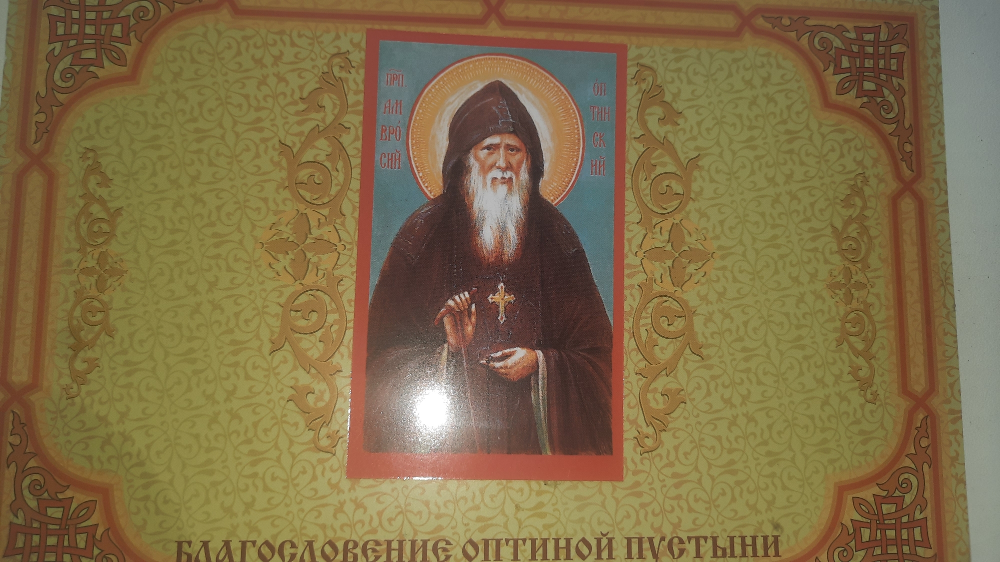 A religious icon depicts a bearded man with a halo, dressed in dark robes. The icon is framed by an ornate border with floral and geometric patterns in gold and orange on a pale yellow background. Vertical text in a Cyrillic script appears on the left side of the image, reading "БЛАГОСЛОВЕНИЕ ОПТИНОЙ ПУСТЫНИ" which translates to "Blessing of Optina Pustyn". The icon and its decorative border appear to be part of a book cover or a devotional item. There is no discernible information within the image that places it specifically in the city of Шклоў, Belarus.
