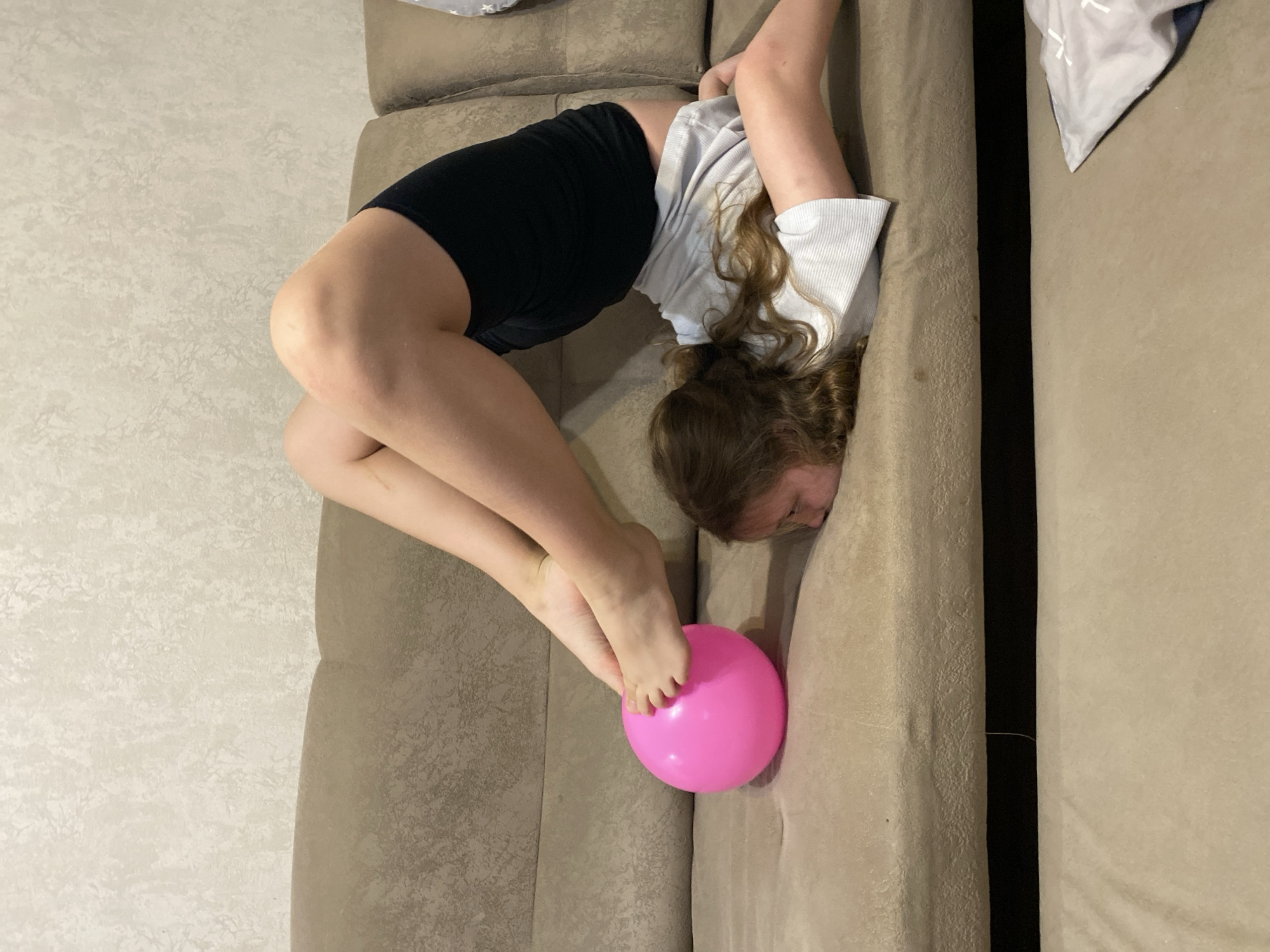A girl is bent over on a beige couch. She is wearing a white shirt and black shorts. Her head and upper body are resting on the couch cushions. Her legs are raised, and her feet are touching a pink ball. The background is a neutral-colored wall. The location is Unknown, Unknown.