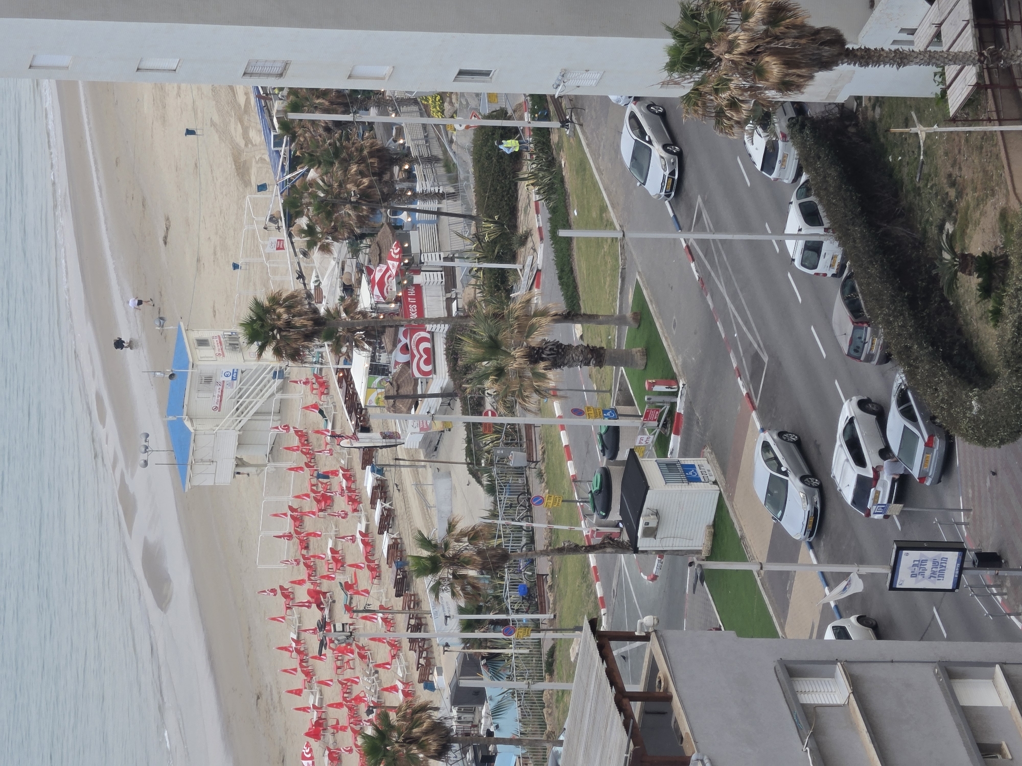 Captured from an elevated perspective in Bat Yam, Israel, the scene is an aerial view of a beach, street, and adjacent buildings. The beach, on the left, is characterized by light-colored sand bordering the water's edge. Numerous red beach chairs and umbrellas are positioned along the sand, suggesting a popular beach destination. Adjacent to the beach is a structure possibly related to beach services, like a lifeguard station. A street runs parallel to the beach, with parked white cars and green grassy areas separating the road from the beach. Palm trees are scattered along the roadside. On the other side of the street, there is a more densely vegetated area. A building on either side of the frame suggests an urban environment. Signage in Hebrew can be spotted along the street. The overall view highlights a seaside urban environment during what appears to be a sunny day.