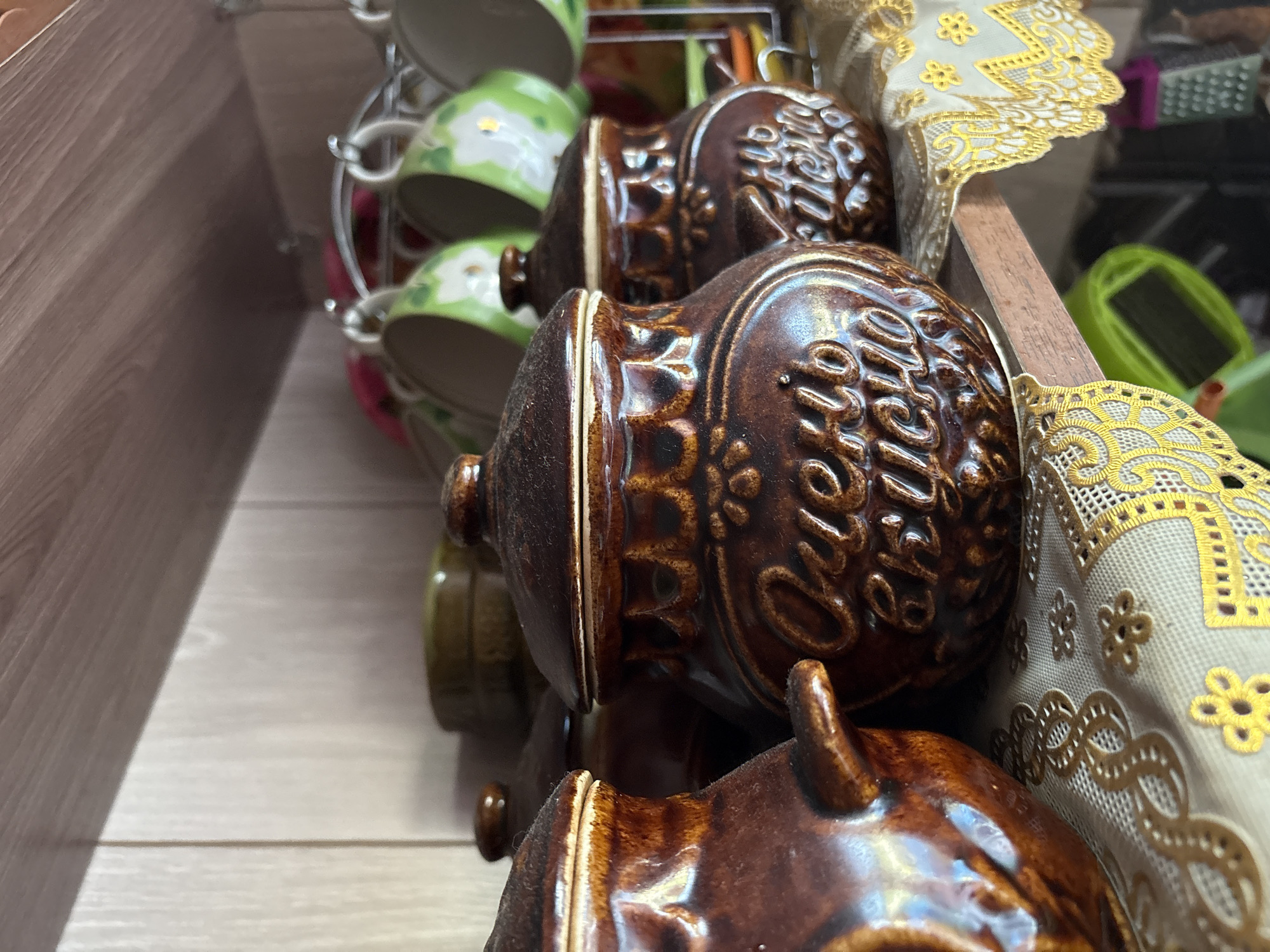 The image shows two brown ceramic pots with lids on a shelf. The pot in the foreground on the right has the words "Очень вкусно" (Very tasty) inscribed on it in Cyrillic letters. The pot on the left is partially visible and appears to have a handle. Behind the pots, several green and white ceramic mugs are hanging from a rack. The shelf is made of wood, and the background consists of a light-colored, wood-paneled wall. A yellow and white decorative fabric with an ornate pattern is draped over the shelf in front of the pots. The scene is domestic, likely a kitchen or pantry. The location of Ветлужский, Russia is not visually evident in the image.