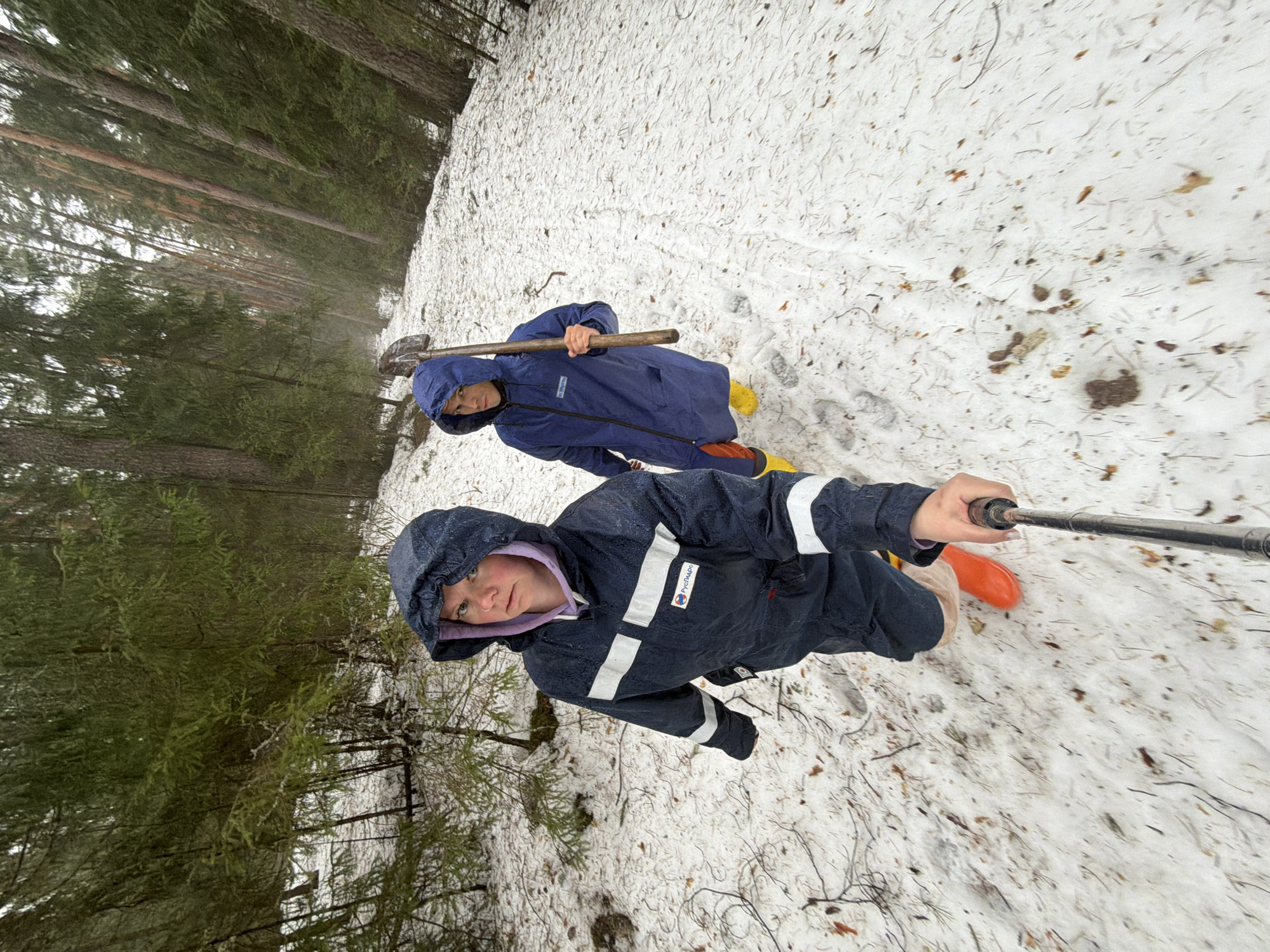 Two individuals are positioned on a snowy terrain bordered by trees, potentially a forest area. The foreground figure, likely a child or teenager, wears a dark hooded jacket with reflective white stripes and holds a selfie stick. Behind them, another person is clad in a blue hooded coat and carries a shovel over their shoulder. They are wearing bright yellow shoes, while the person in front has orange-red boots. The snowy ground is interspersed with fallen leaves and twigs. The perspective suggests the photo was taken from a low angle, likely using the selfie stick held by the foreground person. It is not possible to conclusively determine the location as Chaikovsky, Russia, based on the provided image alone.