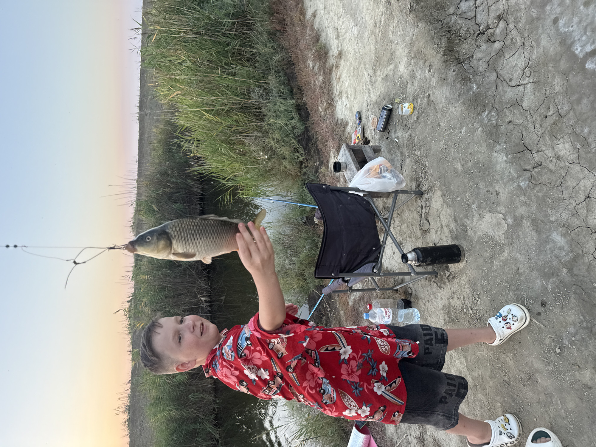 The image depicts a young boy standing in what appears to be a rural setting, possibly near a body of water and reeds, typical of the Урожайновское сельское поселение, Ukraine. The boy is holding up a freshly caught fish, suspended by a fishing line. He is wearing a vibrant red short-sleeved shirt with a tropical floral and vehicle print, dark denim shorts, and white clogs decorated with cartoon characters. In the background, there is a black folding chair, a thermos, two plastic water bottles, and some other items scattered on the ground, including a can and a bag of what looks like chips. The sky in the background suggests it is either early morning or late evening, with a gradient of colors from pale yellow to pink and blue. The ground is dry and cracked, indicating a potentially arid environment.