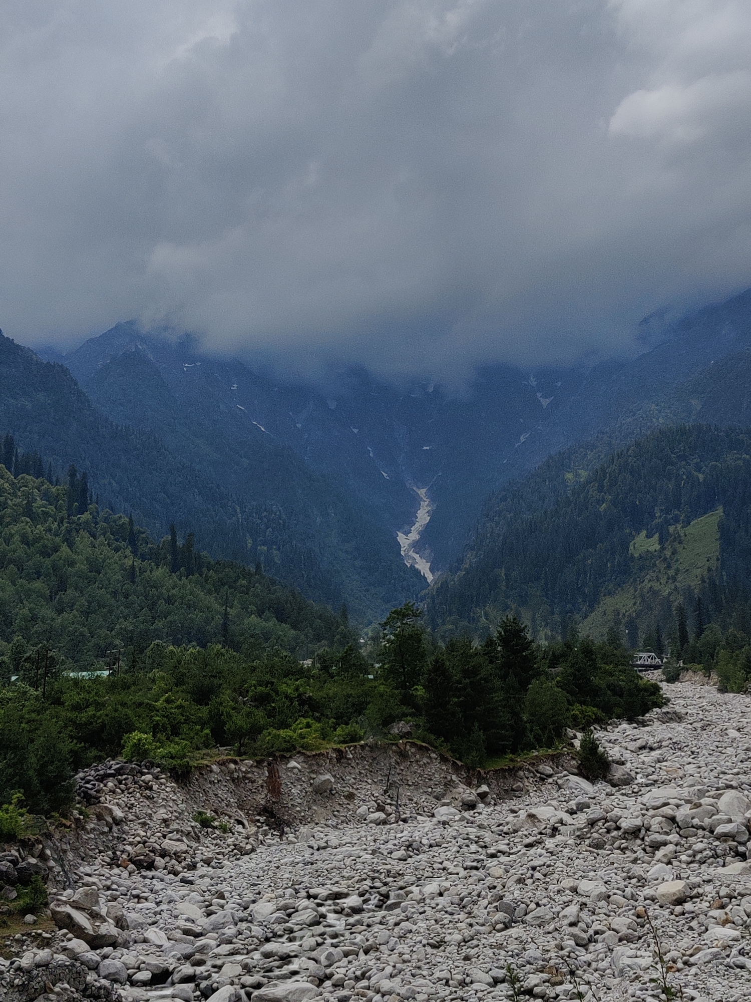 The image depicts a wide, rocky riverbed filled with an abundance of light-colored, smooth stones of varying sizes. A steep, forested slope covered in dense green trees rises on the left side of the frame, leading up to imposing, mist-shrouded mountains in the background. A pale, winding river or stream is visible in the distance, carving its way through a valley between the mountains. The sky is overcast with heavy, dark clouds. On the right side of the image, a faint hint of a bridge structure can be seen amidst the trees. The overall scene suggests a natural, mountainous landscape in Palchan, India, characterized by rugged terrain and lush vegetation.