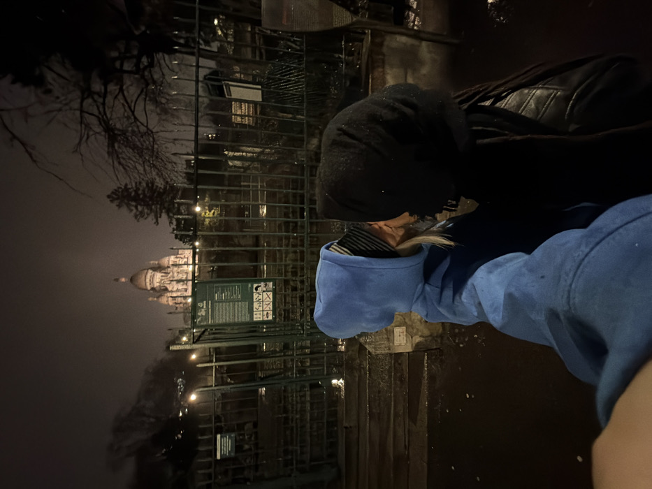 Two hooded figures stand near a wrought iron fence at night. One wears a black hoodie, the other a blue one. Behind the fence, a brightly lit building with domes is visible in the distance. Bare trees are also visible in the background. The ground appears wet, possibly from rain. A sign with symbols is attached to the fence near the figures.