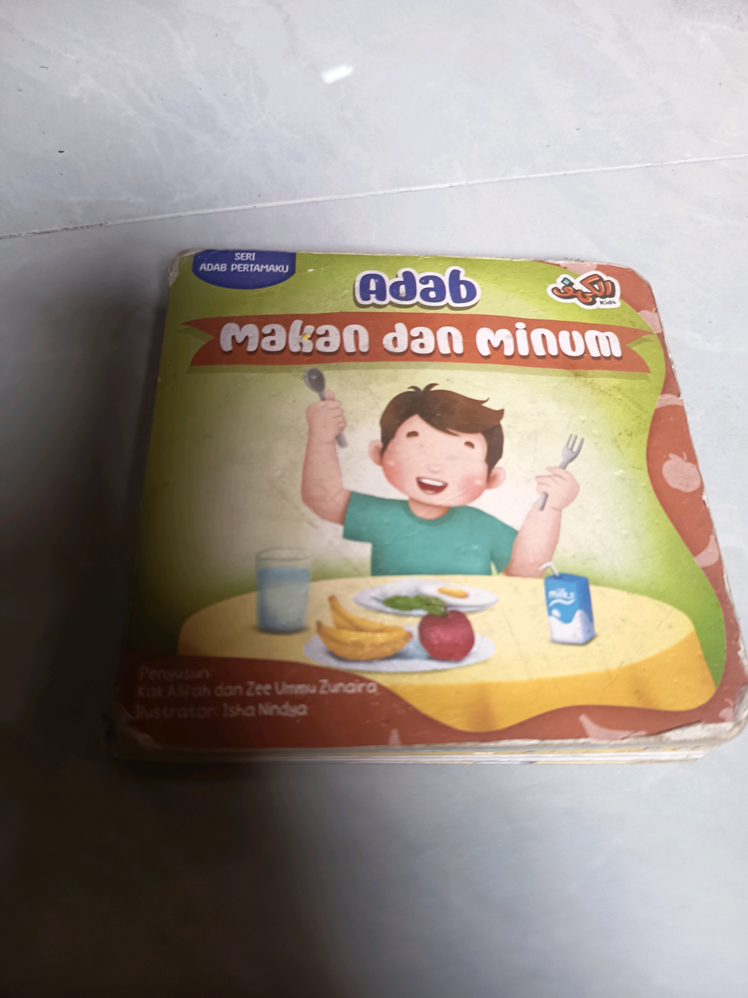 The image shows the cover of a children's book titled "Makan dan Minum" (Eating and Drinking) with illustrations of a young boy eating at a table. The book is part of the "Seri Adab Pertamaku" (My First Manners) series. The boy is holding a spoon in his right hand and a fork in his left. On the table, there are plates with food, a glass of water, and a carton of milk. The background is a light-colored, textured surface, possibly a wall or floor. The book is placed on this surface. No specific location in Sidorejo, Indonesia is depicted or mentioned.