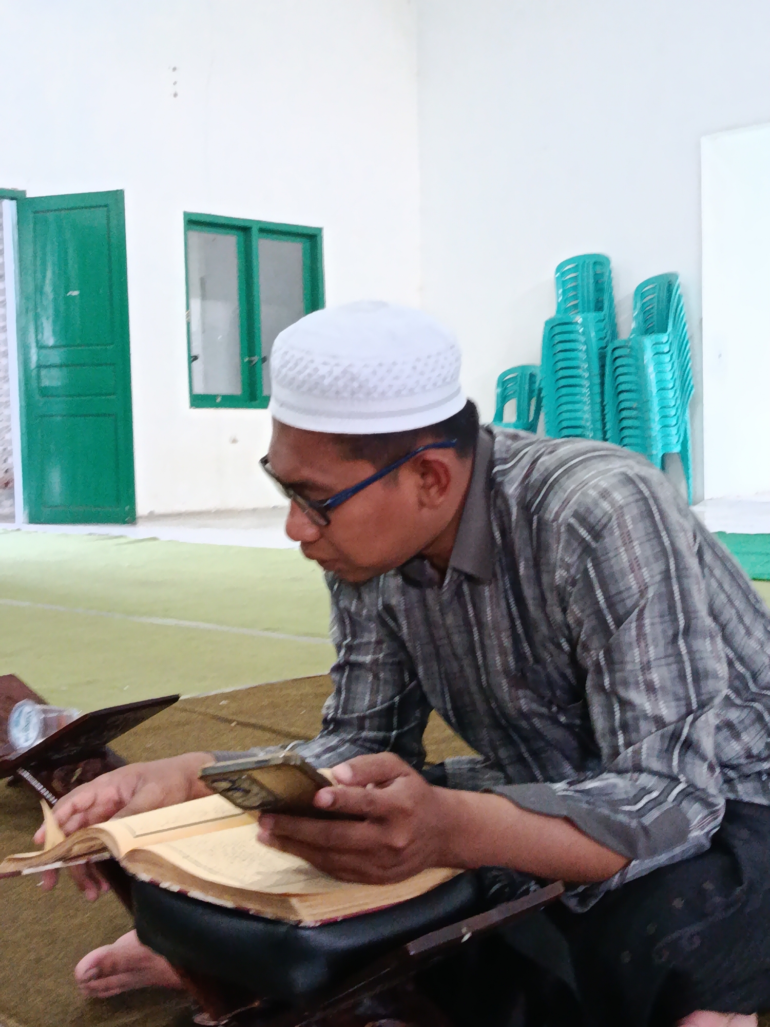A man wearing a white prayer cap and glasses is sitting on a carpeted floor in what appears to be a room with white walls. He is looking down at an open book, which is resting on a wooden stand. His right hand holds a small object, possibly a phone, and his left hand is touching the pages of the book. To his right, a stack of turquoise plastic chairs is visible. In the background, there are two green doors and a window with green frames. The overall environment suggests a public building, possibly a mosque or community center, in Bondowoso, Indonesia.
