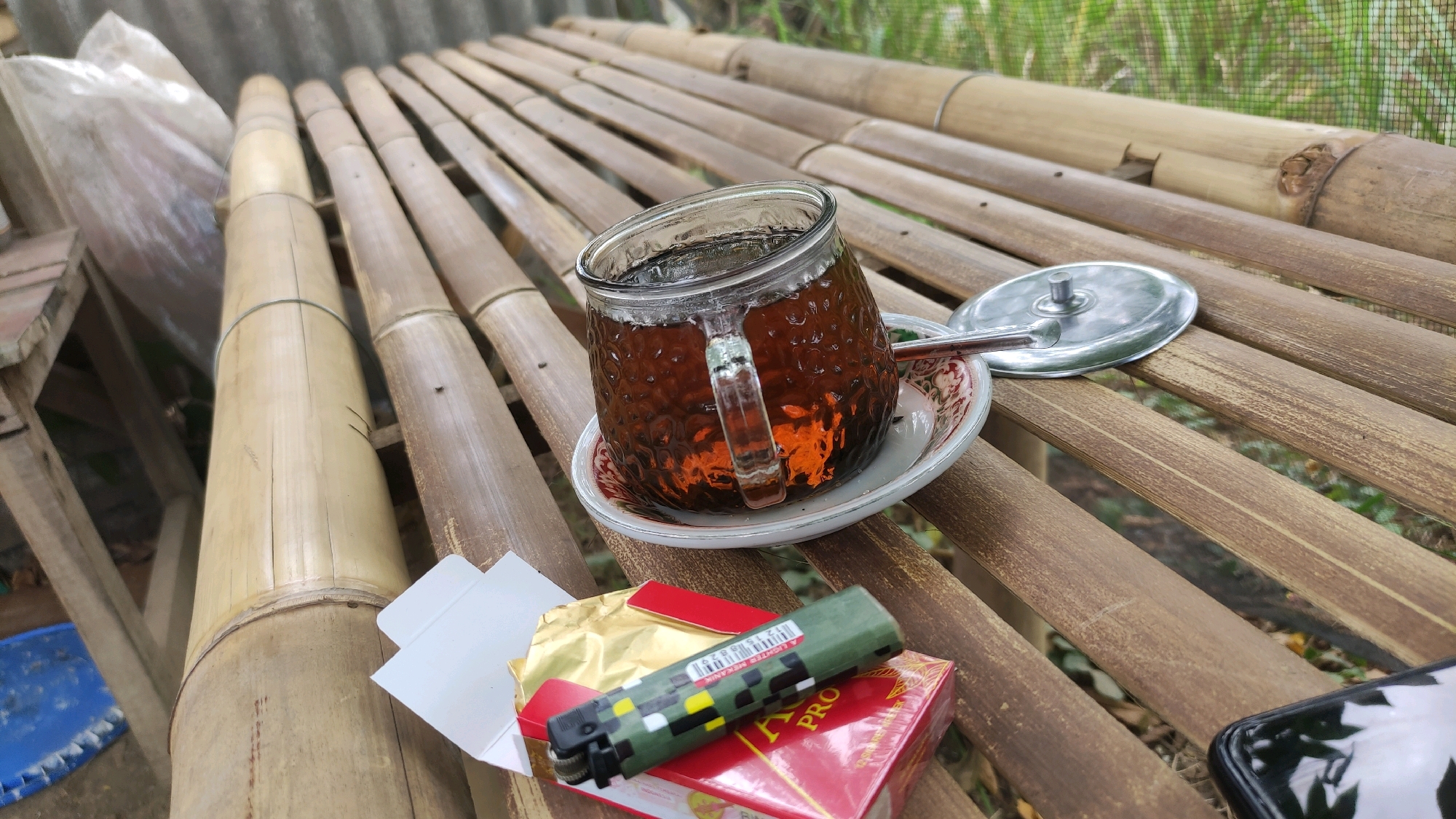 A glass of tea sits on a bamboo table, with a metal lid beside it. In front of the tea, there's a pack of cigarettes, an open packet of gold foil, and a green lighter with a pixelated design. The background shows more bamboo and greenery, suggesting an outdoor setting. The scene appears to be in a rural area, possibly a village like Tiudan, Indonesia, given the prevalence of bamboo structures and natural surroundings.