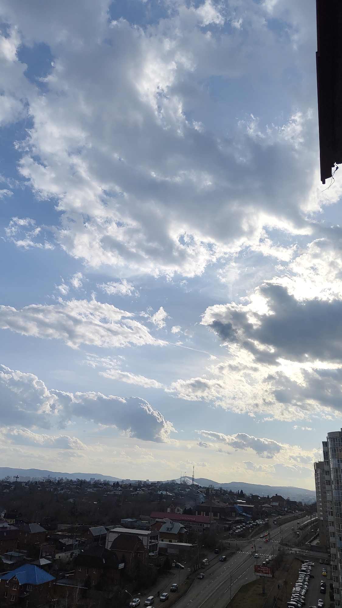 The image shows an elevated view of Krasnoyarsk, Russia under a cloudy sky. Buildings and houses are visible in the midground, leading to a horizon with distant hills or mountains. A multi-lane road with moving vehicles runs through the scene. A tall apartment building is on the right. The sky is predominantly blue with patches of white and gray clouds. A small portion of a building structure is visible in the top right corner.