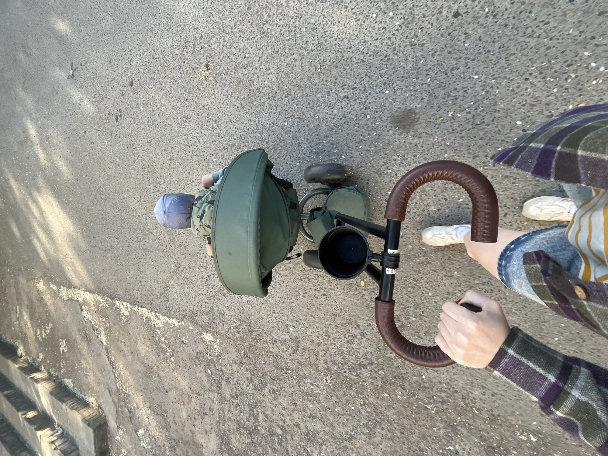 A top-down shot shows a person pushing a child in a green pushchair/stroller on a grey paved surface. The person pushing the stroller wears a plaid shirt, denim shorts, and white sneakers. The child in the stroller wears a cap and is partially obscured by the stroller's hood. The stroller has a brown handlebar, a black cup holder, and a green storage compartment. The paved surface appears to be a sidewalk or pedestrian walkway. The exact location in Chaikovsky, Russia cannot be determined from the image alone.