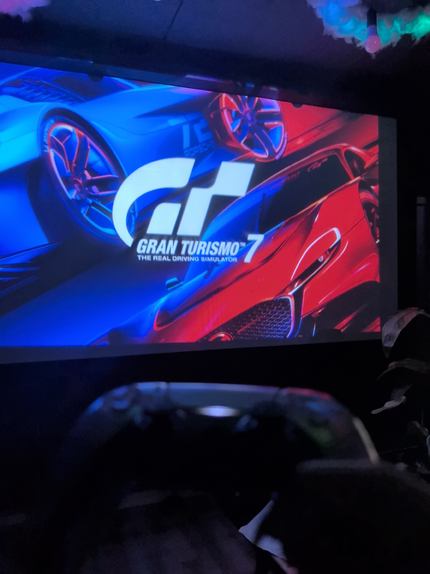 A medium shot depicts a screen displaying the title screen of the video game "Gran Turismo 7." Dominating the background, the screen shows a blue and red sports car. A controller is present in the foreground, partially obscuring the view. Above the screen, a string of decorative lights is visible. The image is taken in a dimly lit room. Location information for Noviy, Russia is not available in the image.