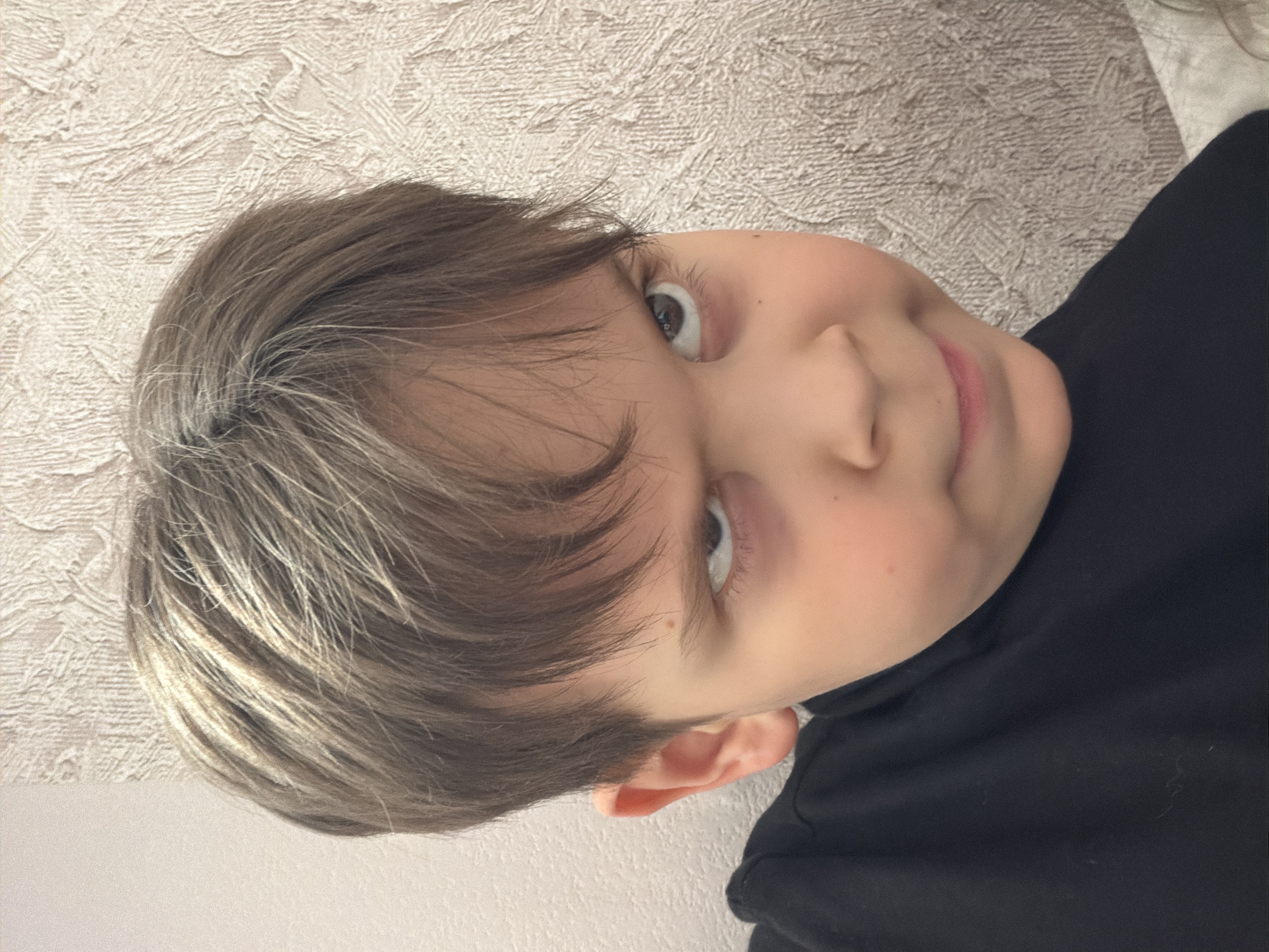 A child with short, light brown and grey hair is depicted in a portrait-style image. The child is wearing a black turtleneck sweater. The background is a textured, off-white wall. The location, Perm, Russia, is not discernible from the image.