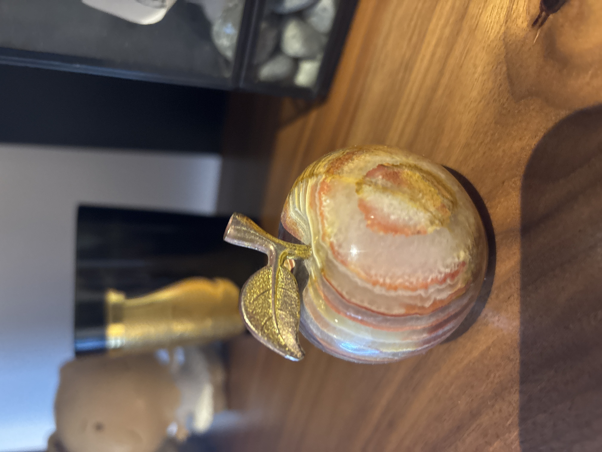 The image shows a close-up, shallow depth-of-field view of a decorative agate apple figurine placed on a wooden surface. The apple is round with distinct banding of white, pink, and yellow, and features a golden leaf and stem. In the blurred background, a gold-colored vase and a black cylindrical object are visible. To the right, a glass display case containing gray stones is partially visible. The setting appears to be indoors, on a table or shelf, and does not provide any specific information about its location in Dubai, United Arab Emirates.