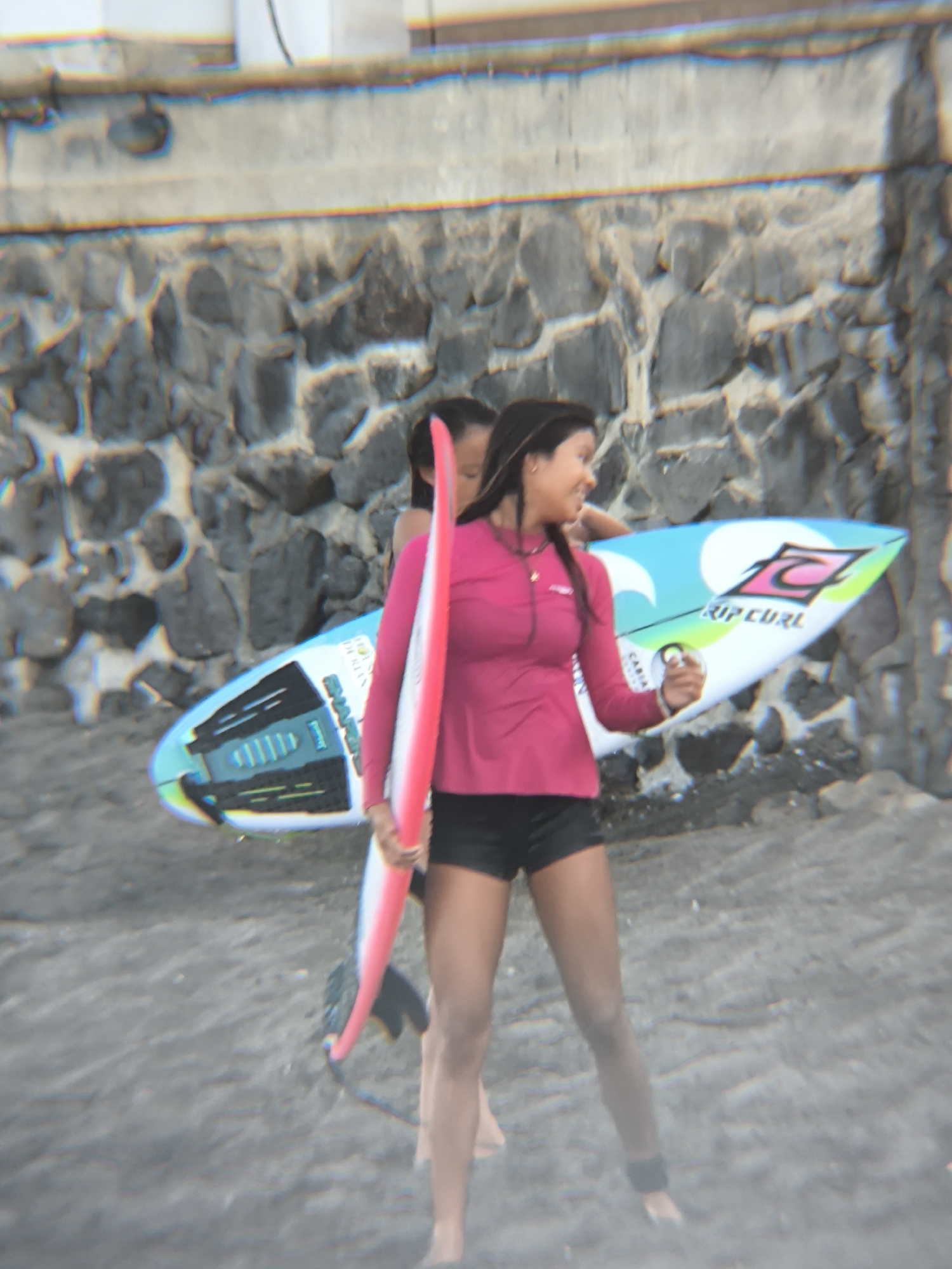 In North Kuta, Indonesia, two individuals stand on a sandy beach, their figures partially obscured by a light haze. The woman in the foreground wears a long-sleeved pink rash guard and black shorts. She holds a pink and white surfboard vertically at her side. A second individual stands behind her, partially visible, holding a larger, colorful surfboard with the text "RIP CURL" on it. A stone wall serves as the backdrop, with a lighter-colored building above it.