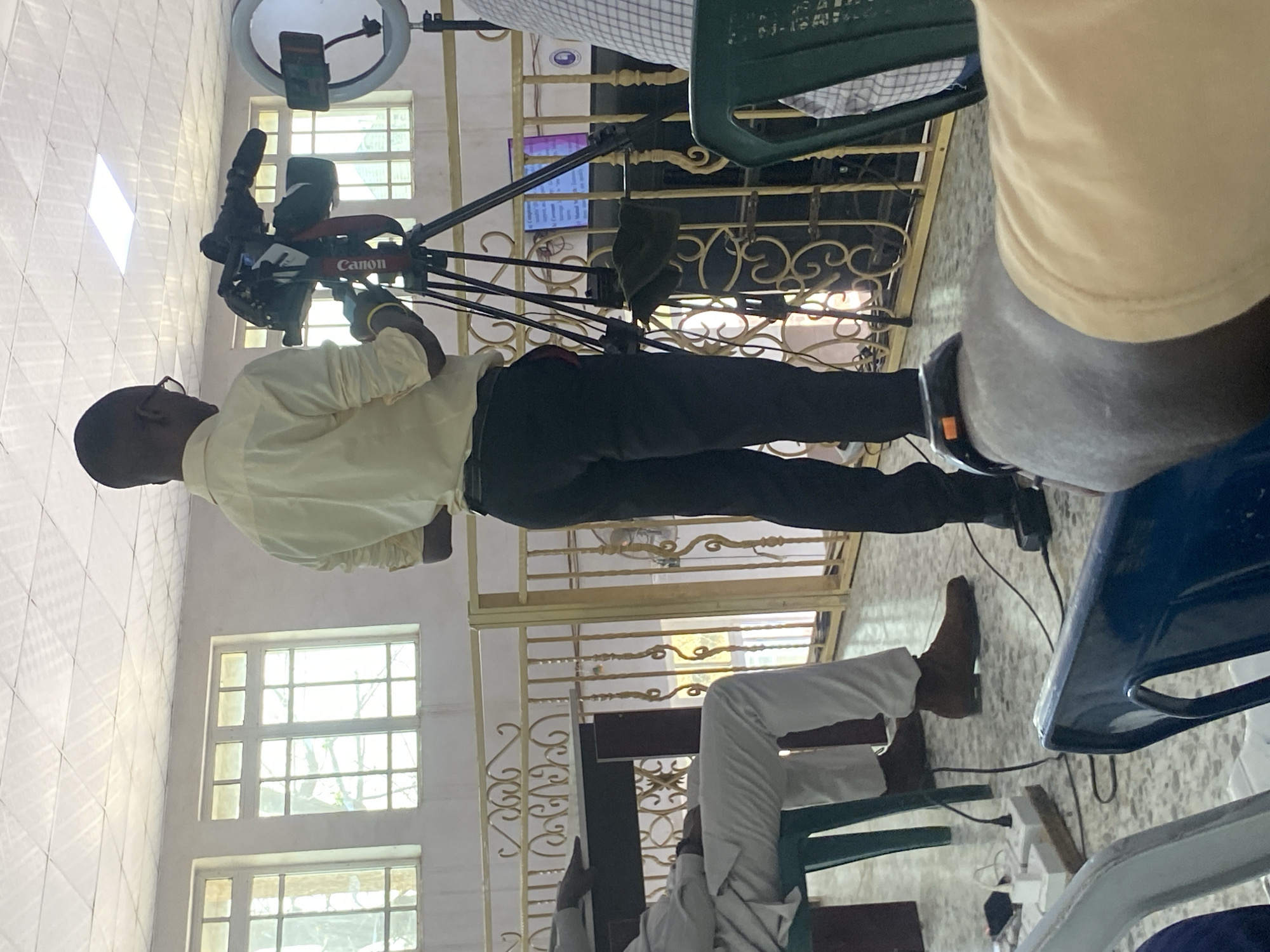 A man in a light yellow shirt and dark pants is operating a Canon video camera on a tripod indoors. The camera is pointed towards a screen displaying text. The location appears to be a well-lit building with multiple windows and white checkered tile flooring. The picture appears to be taken in Barden Yana, Nigeria, although not evident from the image itself. Other people are present, including one wearing white pants and brown shoes seated on a chair. A green chair and a blue chair are also visible in the foreground.