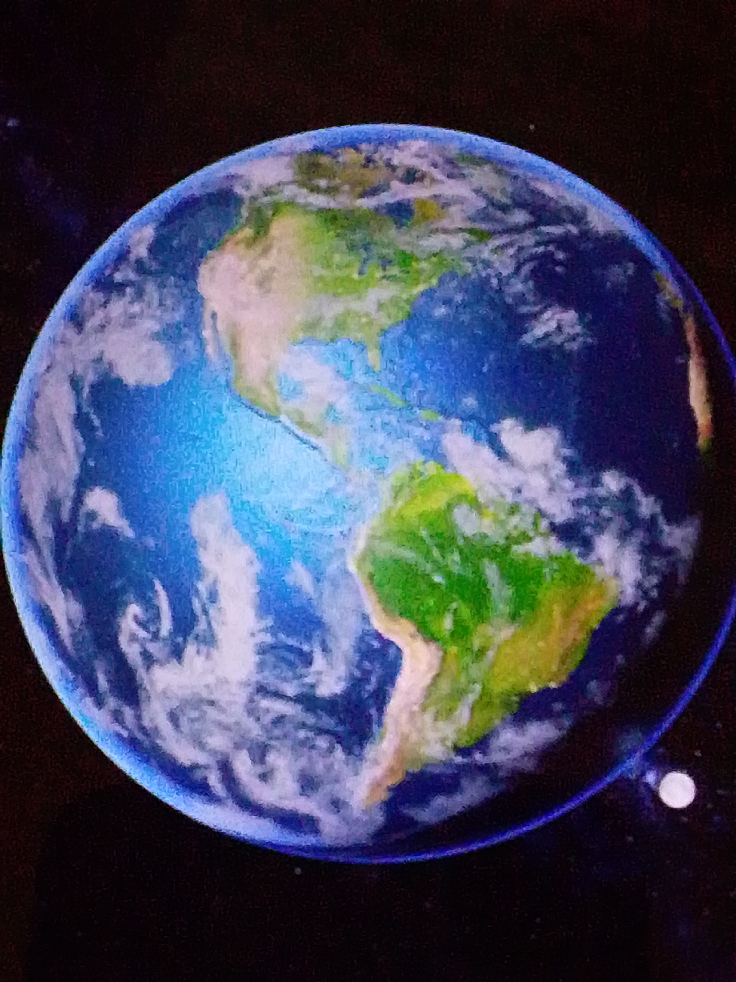 The image shows a view of the Earth from space, with a portion of North and South America visible, along with clouds and oceans. The Earth is set against a dark background representing space, with small white dots that could be stars or distant celestial bodies. In the lower right portion of the image, a small, pale orb is visible, likely representing the Moon. The rendering of the Earth appears to be stylized, possibly from a painting or illustration, with visible brushstrokes. There are no people, buildings, or any elements that would indicate a specific location within the city of Aznakayevo, Russia, or any other city. The scene is entirely celestial.
