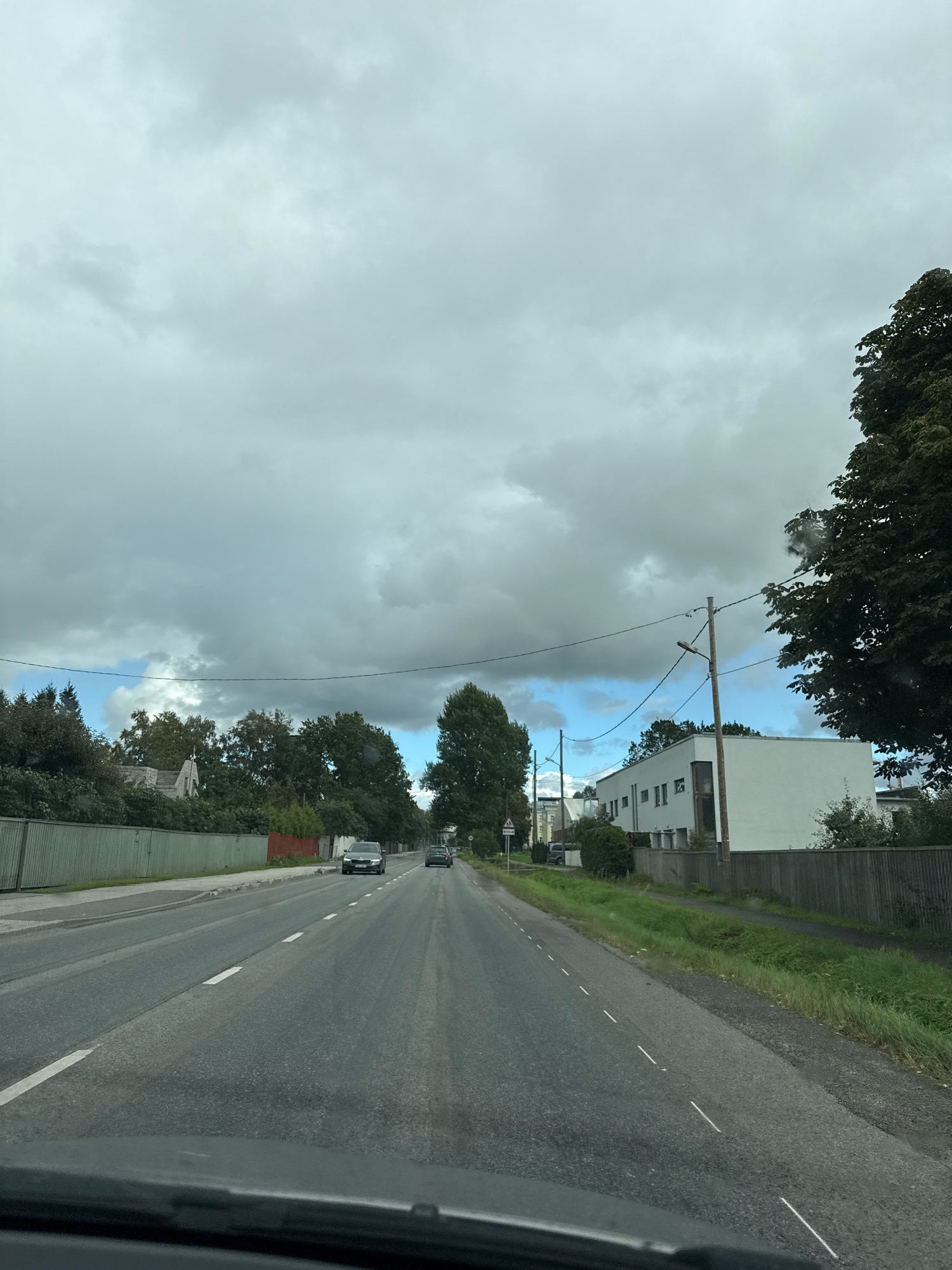 The image captures a view from inside a vehicle traveling on a road in Tallinn, Estonia. The sky is overcast with dark, heavy clouds. On the left side of the road, there is a long, green fence and trees lining the property behind it. Further down the road, a building with a light-colored roof and grey walls is visible. On the right side of the road, a modern, white, rectangular building with multiple windows is situated behind a wooden fence. There is a power line running across the scene. The road itself is asphalt with white dashed lines marking the lanes. Two cars are visible on the road, traveling in the same direction as the vehicle from which the photo is taken. The environment suggests a suburban or semi-urban setting.
