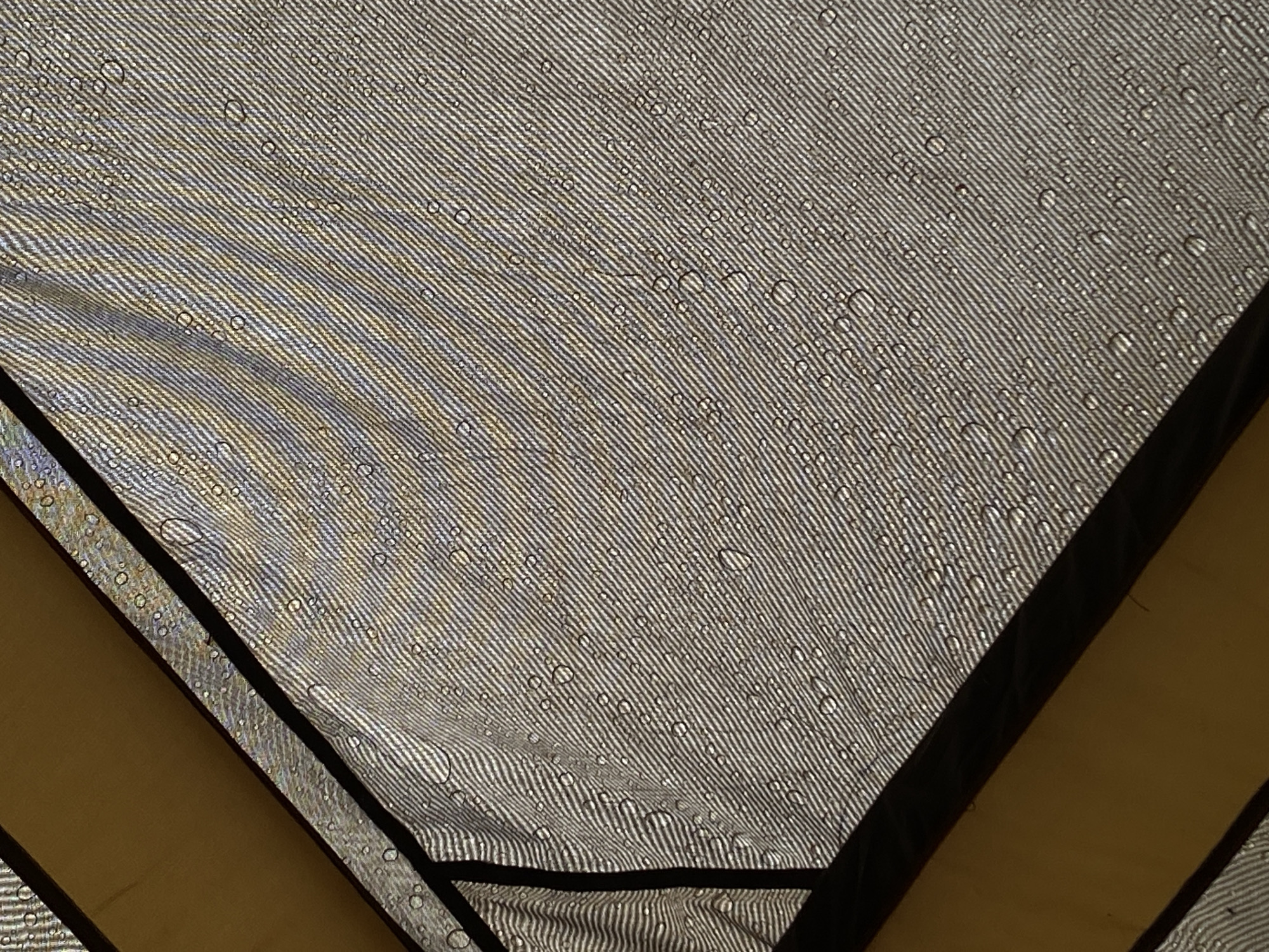 An upward-facing angled shot shows a section of a light-colored tarp with water droplets on its surface. The tarp is textured with a fine, ribbed pattern and appears to be stretched taut, supported by dark framing that forms sharp diagonal lines across the image. A sliver of a yellow surface is visible in the upper left and lower left corners, appearing to be another section of the structure supporting the tarp. The water droplets are of varying sizes, some forming small beads and others larger, irregular shapes, reflecting the light and the surrounding environment. The scene is outdoors, suggested by the lighting and the presence of water on an exterior covering. This image does not depict any specific location in Baltiiskoe, Russia, or any people.