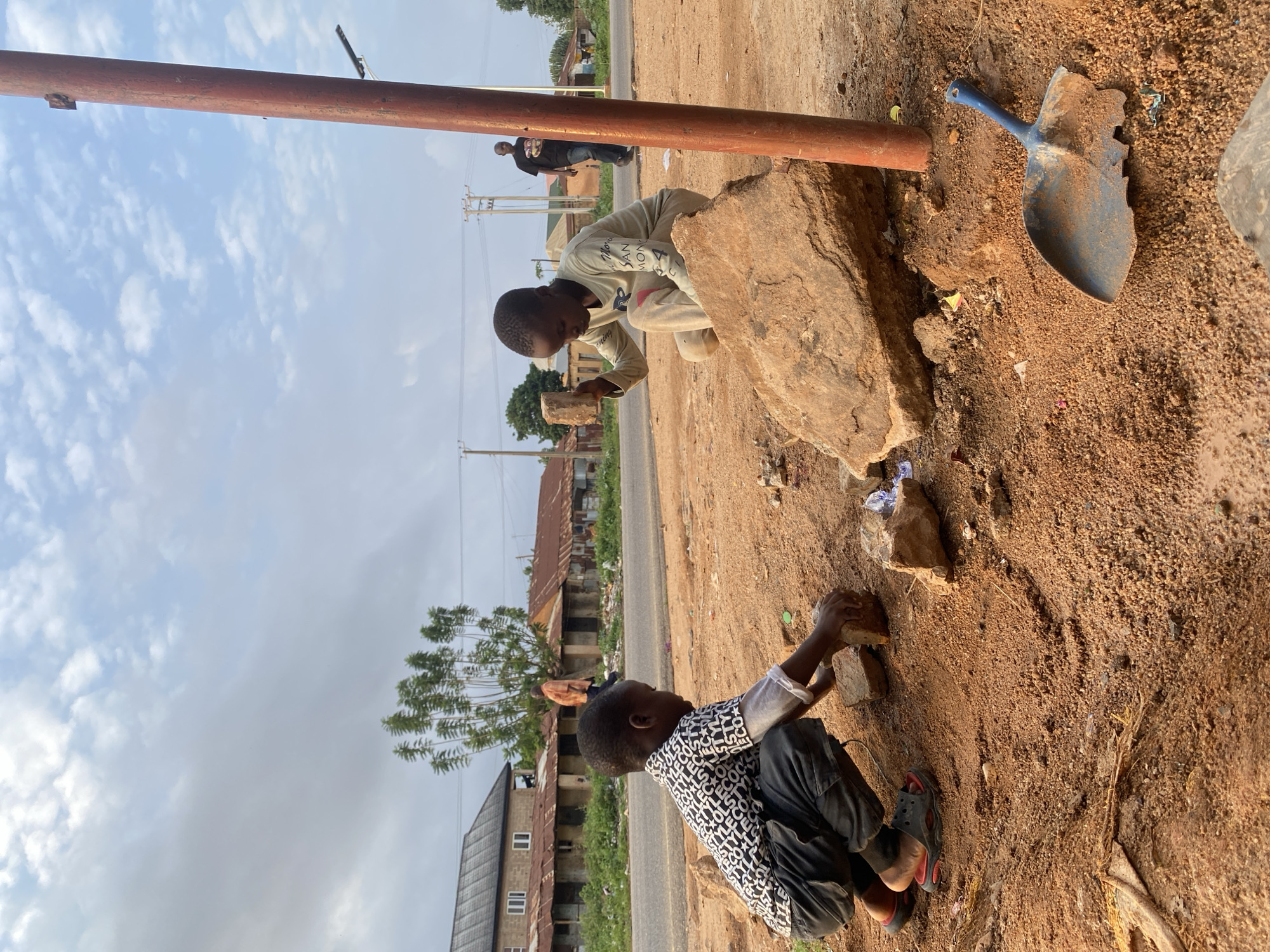 Two young boys are playing near a road in Kuru Dungus, Nigeria. One boy is crouched, wearing a black-and-white patterned shirt and dark pants, holding a small brick or stone. The second boy is kneeling near a large rock, wearing a light-colored shirt. A rusty metal pole stands vertically next to the boys. A blue, heavily used shovel rests on the ground near the rock. In the background, there is a road, buildings with corrugated metal roofs, and a person walking. The ground is sandy and littered with debris.