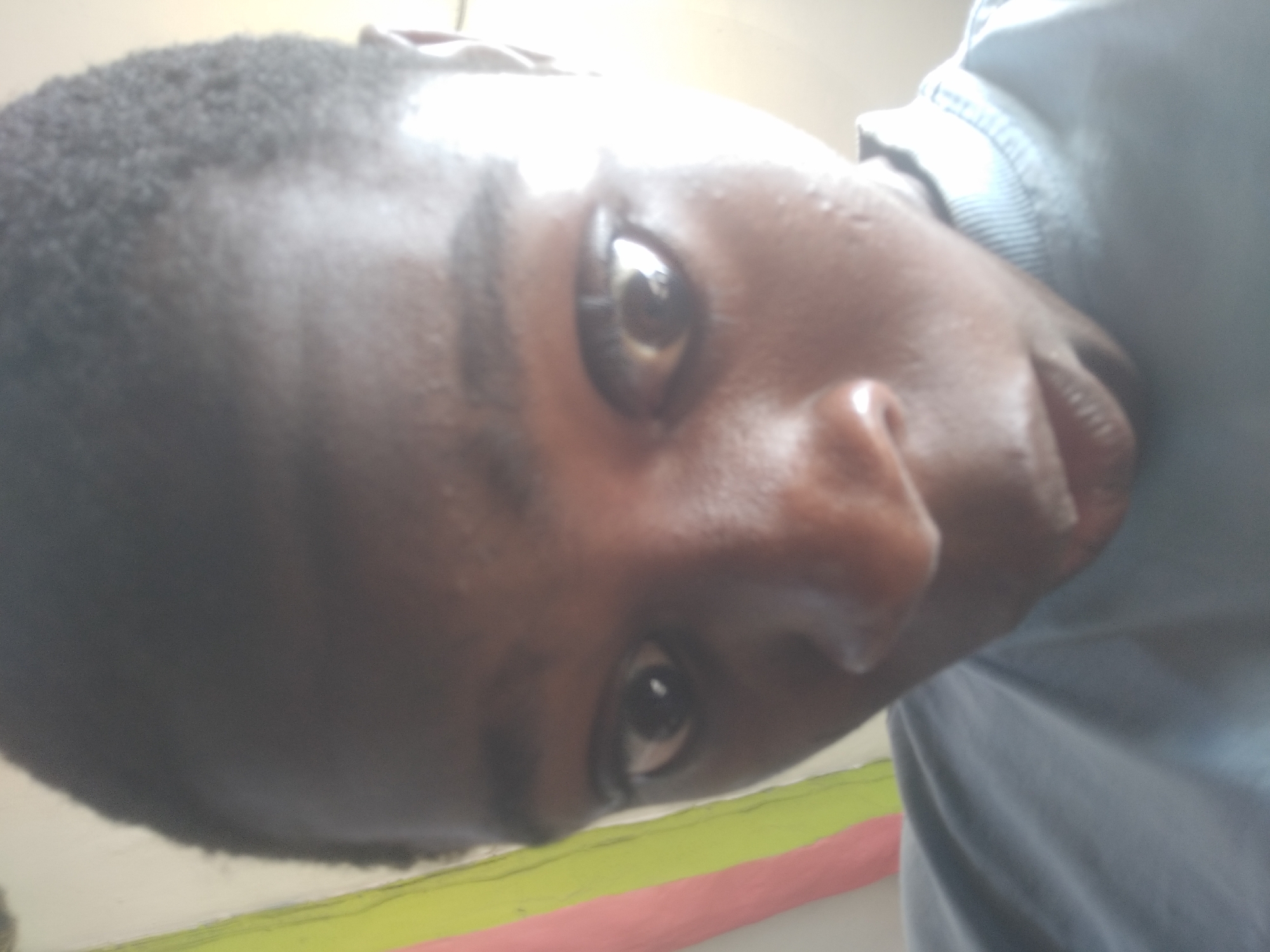 A close-up photograph depicts a person with dark skin and short, dark hair. The person's face is the main subject, showing details of their eyes, nose, and mouth. The person is wearing a light blue shirt. The background is blurry but appears to show a wall with colored stripes. The image angle is rotated. Location in Shangisha, Nigeria cannot be determined from the image.