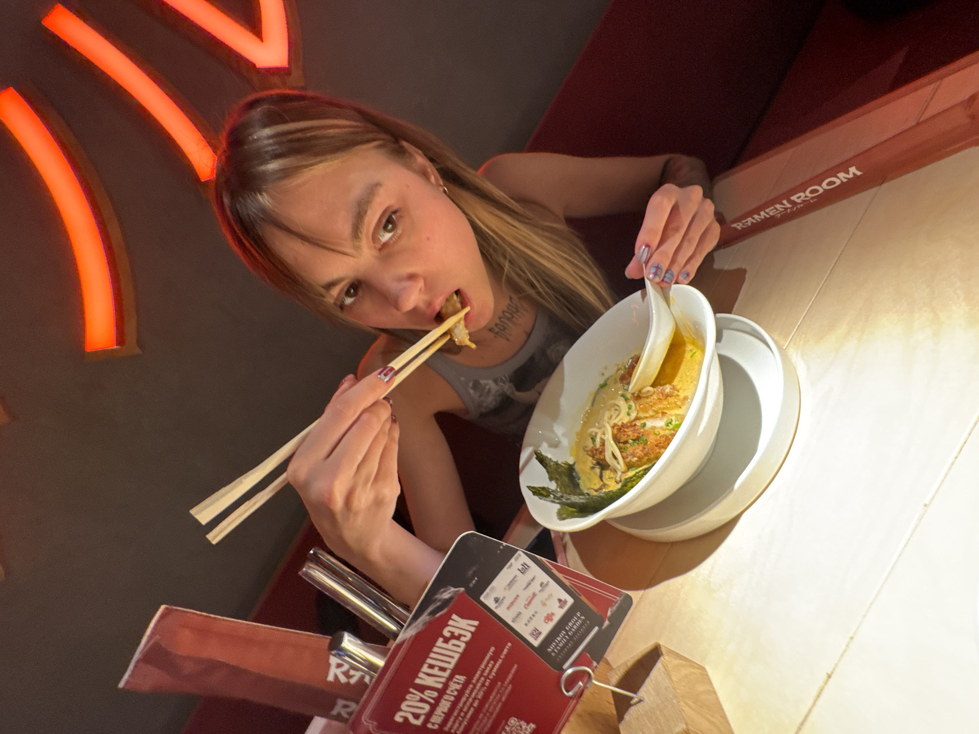 The image shows a young woman with blonde hair and bangs sitting at a table in a dimly lit restaurant. She is holding chopsticks and eating from a bowl of ramen. The restaurant appears to be decorated with red neon lights on the ceiling. On the table, there is a red promotional card with Russian text and logos. The text on the card says "20% КЕШБЭК С ПЕРВОГО СЧЕТА", which translates to "20% Cashback from the first bill". The card also shows a smartphone screen with various logos, including "Ramen Room". The woman is wearing a grey tank top. She has tattoos on her neck. The table surface is light-colored and appears to be made of wood. The background is dark, with red seating visible on the right. The overall ambiance suggests a modern and casual dining establishment, likely a ramen restaurant. The presence of the "Ramen Room" logo on the promotional material indicates the establishment's name. Given the Russian text, the location is highly likely to be within Russia. The presence of "Ramen Room" in Cyrillic script on the promotional material, alongside English text "RAMEN ROOM" on the red menu stand, suggests that this restaurant may be located in Moscow, Russia, a major city with international dining establishments.