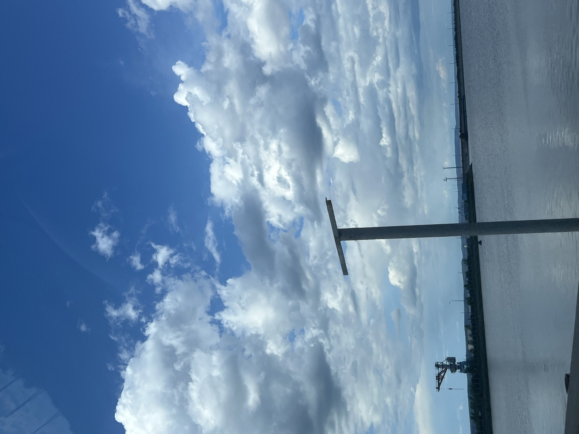 The image shows a cloudy sky over a body of water, possibly a river or reservoir, with a visible shoreline in the distance. A metal structure, possibly a bridge or pier support, is in the foreground. There is a crane near the shore in the middle right, and a series of power lines/electrical towers are visible along the shoreline. The sky is blue with large, white cumulus clouds. The photo may have been taken from a vehicle due to the reflections on the bottom right. The location, Chaikovsky, Russia, is consistent with the presence of large industrial infrastructure and the type of waterways seen.