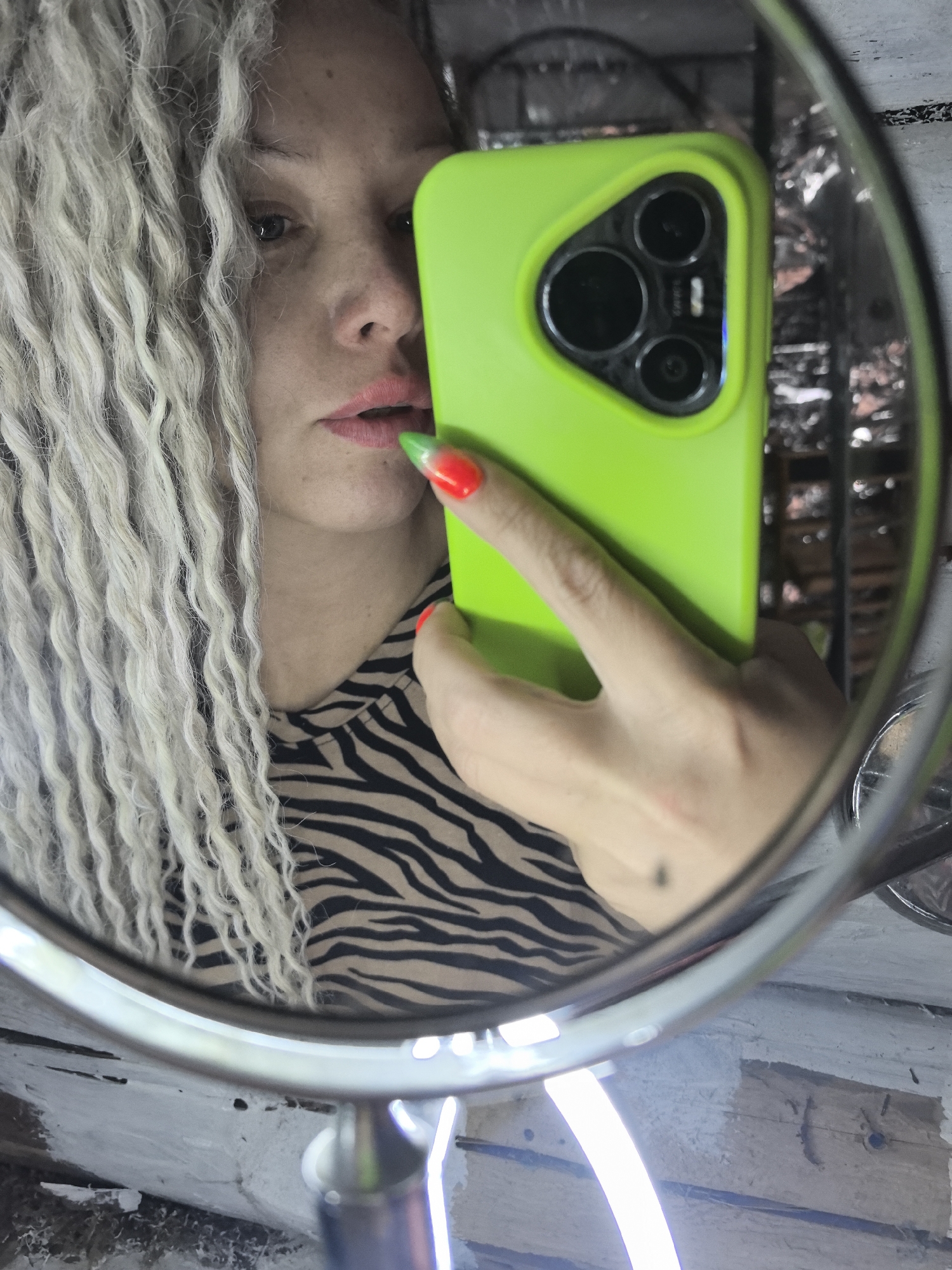 A woman is taking a mirror selfie, holding a phone with a bright green case. Her hair is styled in long, light-colored dreadlocks. She's wearing a zebra-print top. The phone's camera lens arrangement is visible. Her nails are painted with a green-to-red gradient. The background seen in the mirror reflection appears to be an outdoor or semi-outdoor setting with some metallic or wire-like elements and weathered wood or wall. The location is not identifiable as Noviy, Russia.