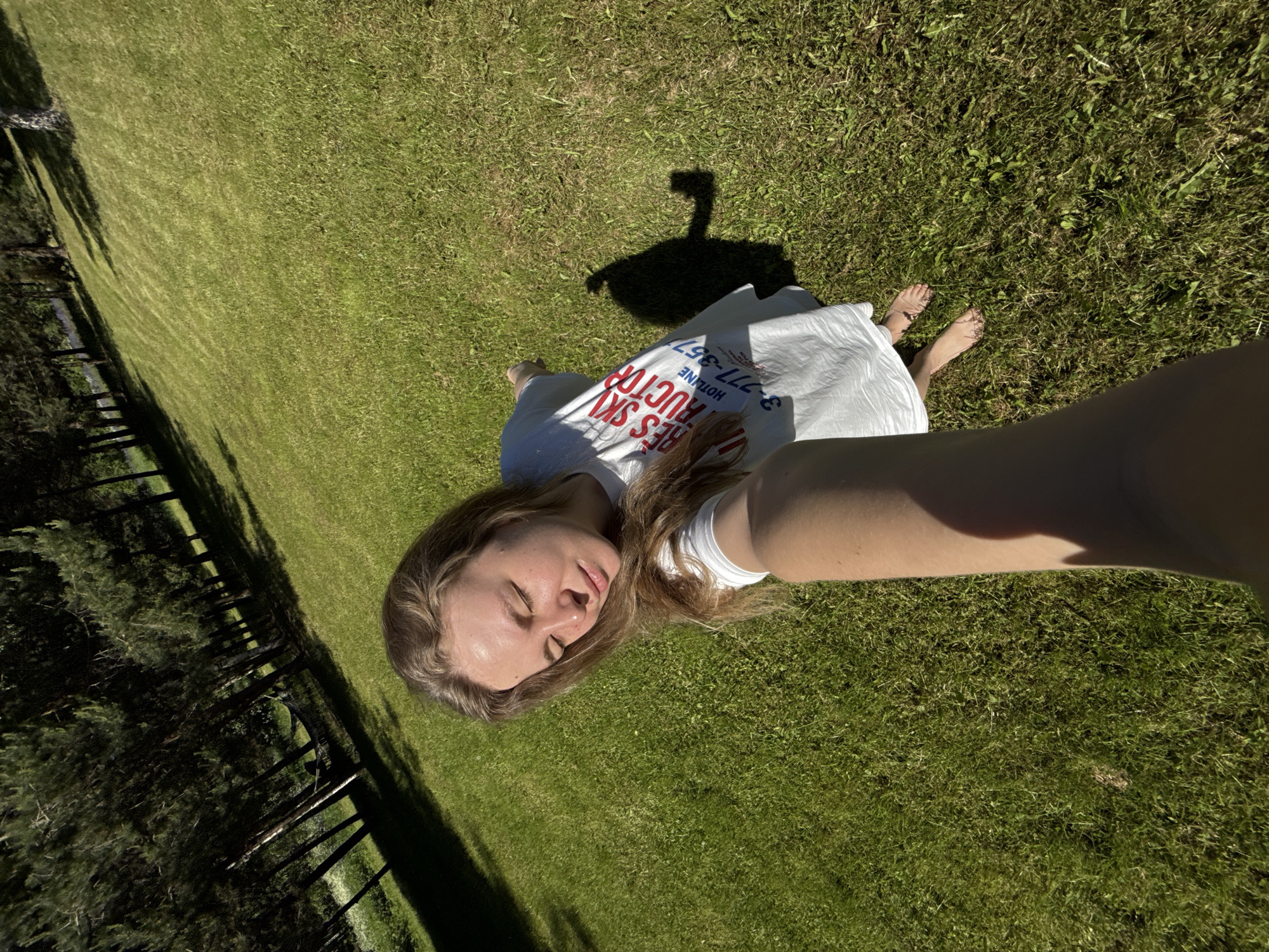 An adult female is captured in a high-angle selfie in an outdoor grassy environment in Копнино, Russia. She has long, light brown hair and is wearing a white t-shirt with "SKI INSTRUCTOR" printed in red text and a partial phone number "3-777-357" in blue text below it. Her eyes are closed, and her face is tilted upwards, indicative of enjoying sunlight. She is barefoot, standing on green grass, which shows some patches of weeds. Her right arm is extended, holding the camera/phone. A pronounced shadow of her body, including the device, is cast on the grass to her right, indicating strong direct sunlight. In the background, a dense line of tall pine trees is visible, with a hammock strung between two of them. The lighting suggests either late morning or late afternoon.