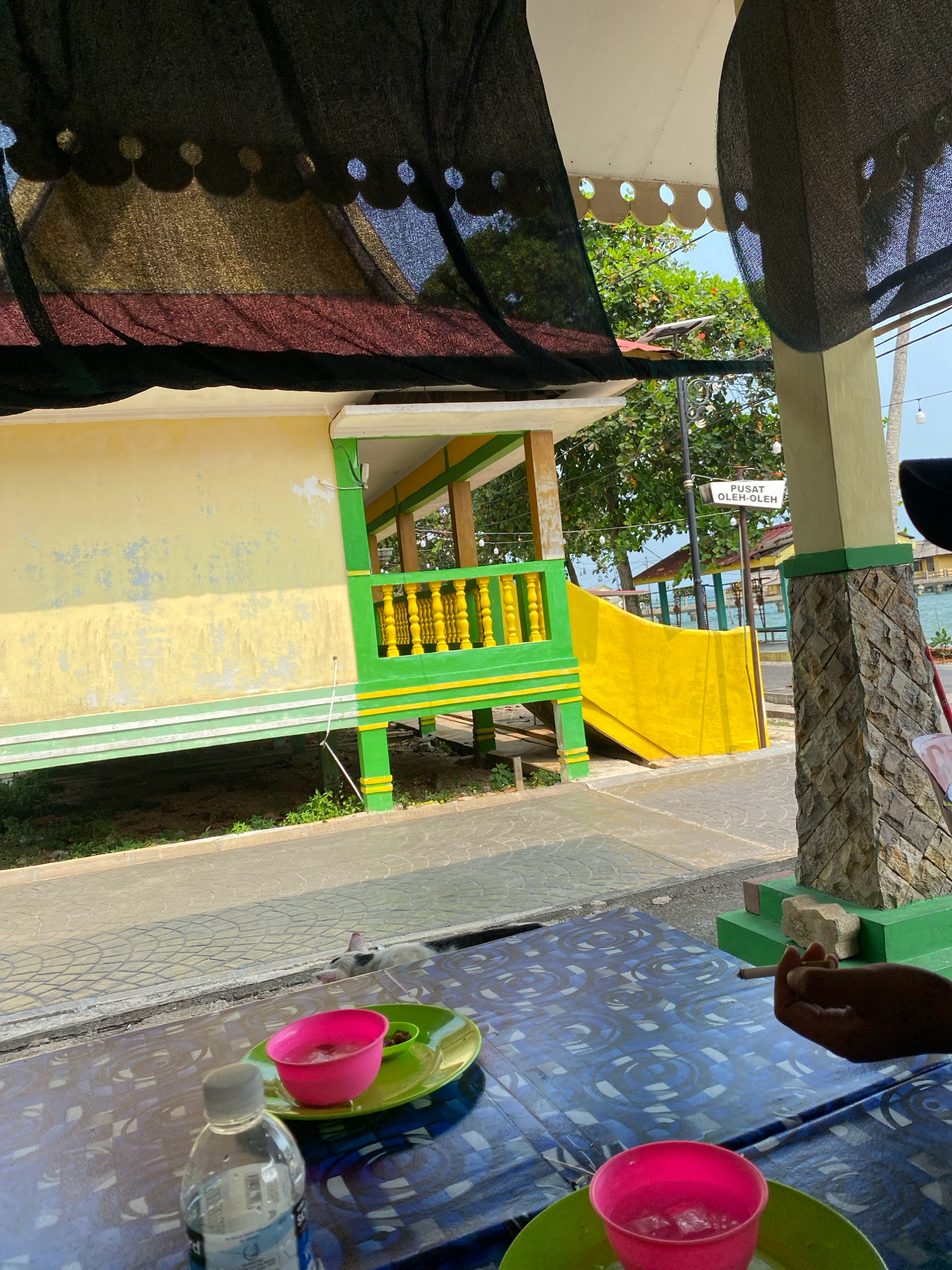 In Tanjung Pinang, Indonesia, a scene unfolds at an outdoor seating area. A table covered with a blue and white patterned cloth is in the foreground, on which rests a water bottle, two green plates with pink cups filled with ice and pink liquid, and a small dish with what appears to be food. A hand is visible, holding a lit cigarette and appearing to smoke. To the left, a white pigeon stands on the patterned ground.
Behind the table, a yellow building with green trim stands on stilts, with a yellow ramp leading up to a green railing. The building appears to be a shop or cafe, indicated by a sign that reads "PUSAT OLEH-OLEH," which translates to "Souvenir Center." Trees and the ocean are visible in the background. Above, a black canopy casts shadows over the scene, suggesting it provides shade for the seating area. The overall impression is a casual, tropical setting.