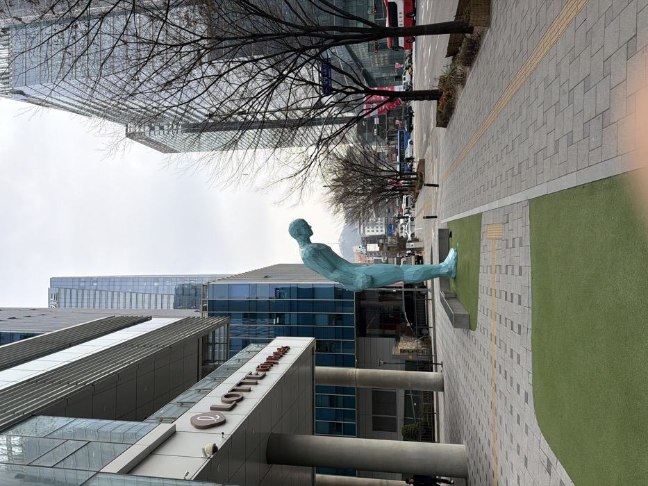 The image displays an urban street scene in front of the Lotte City Hotel Myeongdong, located in Seoul, South Korea. The central subject is a large, teal-colored polygonal sculpture depicting a male figure in a deep bow, positioned on a patch of artificial green turf next to a concrete planter. To the left, the modern glass and concrete facade of the Lotte City Hotel Myeongdong is visible, featuring its branding in red text. A single individual is partially visible walking near the hotel's entrance pillars. To the right, a wide paved sidewalk, featuring grey rectangular pavers and a yellow tactile strip, is lined with bare deciduous trees. Beyond the sidewalk is a multi-lane asphalt road with moderate vehicle traffic, including several cars and red public buses. In the background, numerous tall, modern glass skyscrapers dominate the cityscape, and a distant mountain range is faintly visible under an overcast sky.