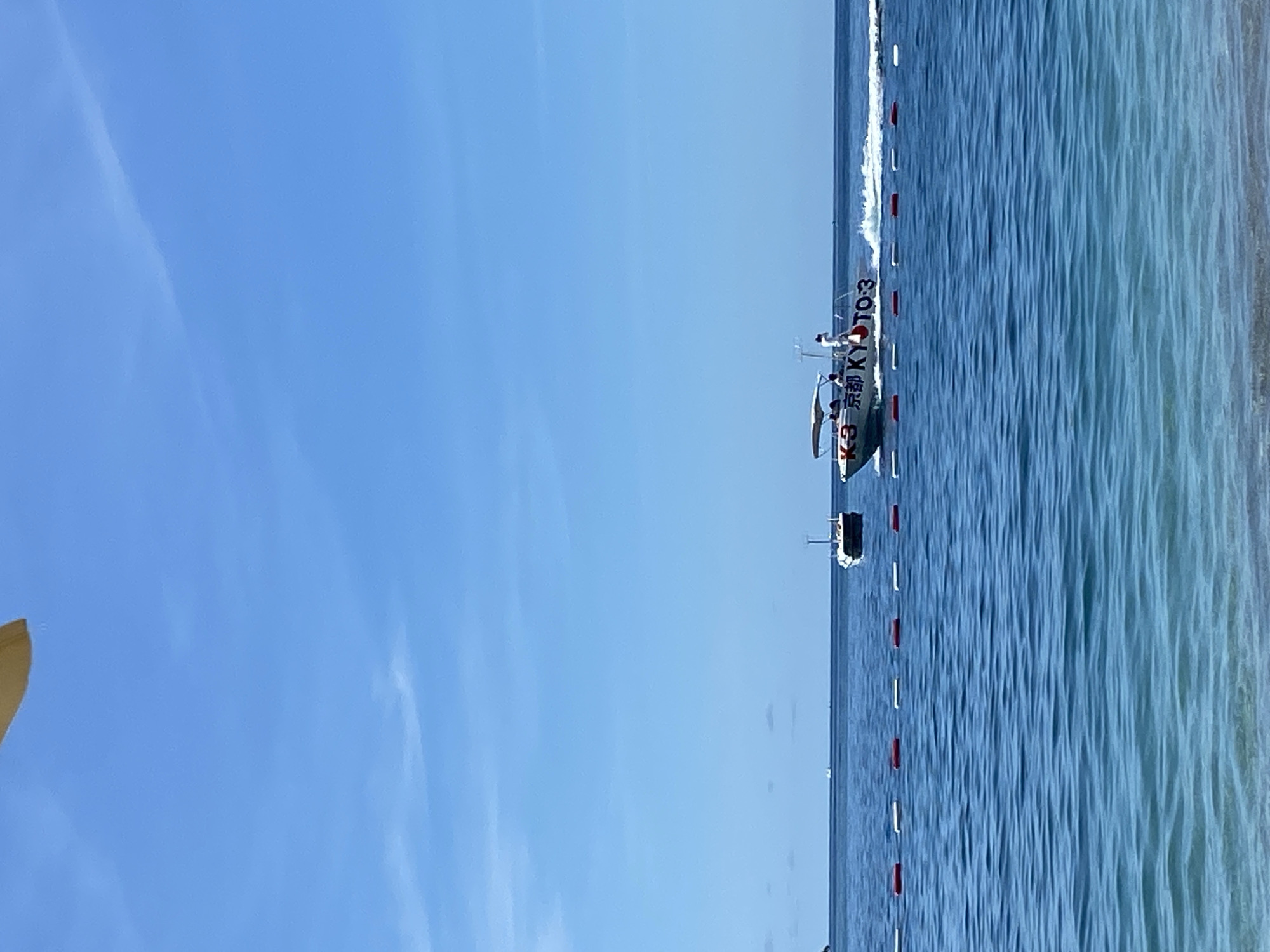 The image depicts a serene daytime scene over a calm blue-green sea, likely in Budva, Montenegro, under a clear, expansive blue sky with minimal cloud cover. A distinct horizon line separates the sky from the water.
The sea features subtle ripples and is marked by several lines of red and white buoys extending into the distance, possibly indicating a boating lane or a designated swimming area. A white boat is the primary subject on the water, actively moving and creating a white wake behind it. Prominently displayed on its side in red lettering are the characters "K-3" and "京都 KYOTO-3", with "京都" being the Japanese name for Kyoto. A few individuals are visible on board the boat, with one person in a white top standing towards the stern. Further along the buoy line, another smaller, dark boat is visible in the distance.
The weather is bright and sunny, indicating a clear day. A blurred, golden-brown object, possibly part of a beach umbrella or similar structure, is partially visible in the bottom-left corner, suggesting a nearby shoreline or beach. The overall scene conveys a sense of calm and leisure activity on the water.